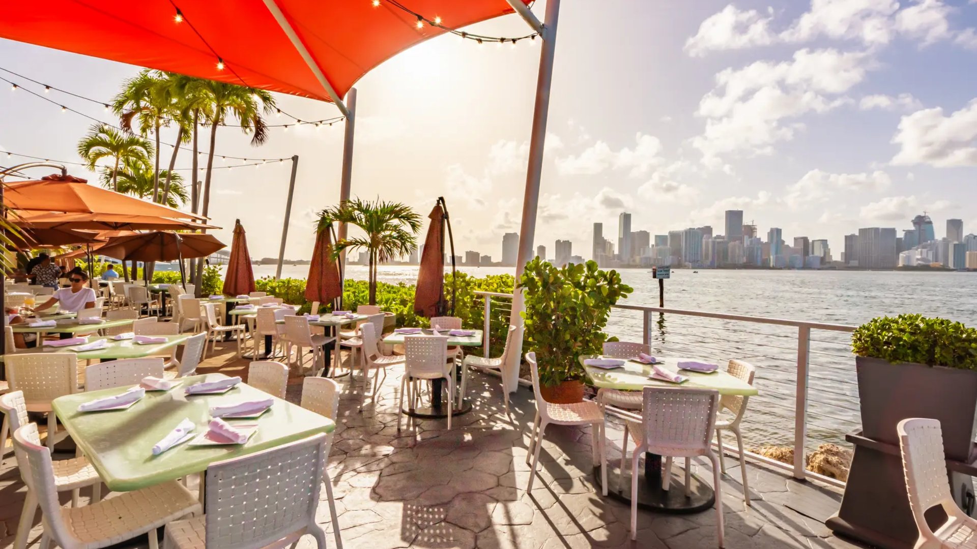 Outdoor waterfront restaurant patio with Miami skyline views and palm trees by The Edit Real Estate