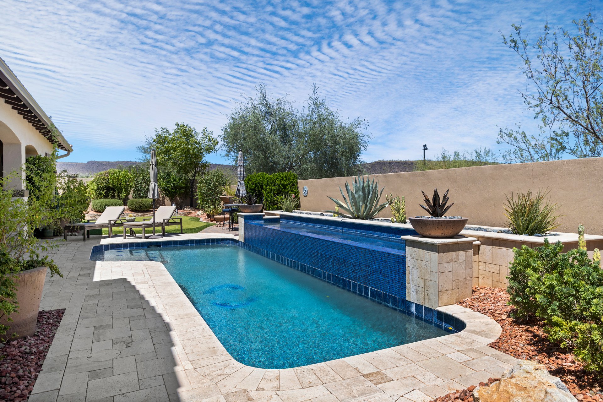 Luxury backyard patio with a narrow blue-tiled pool, lounge chairs, desert landscaping, and mountain views under a bright sky.