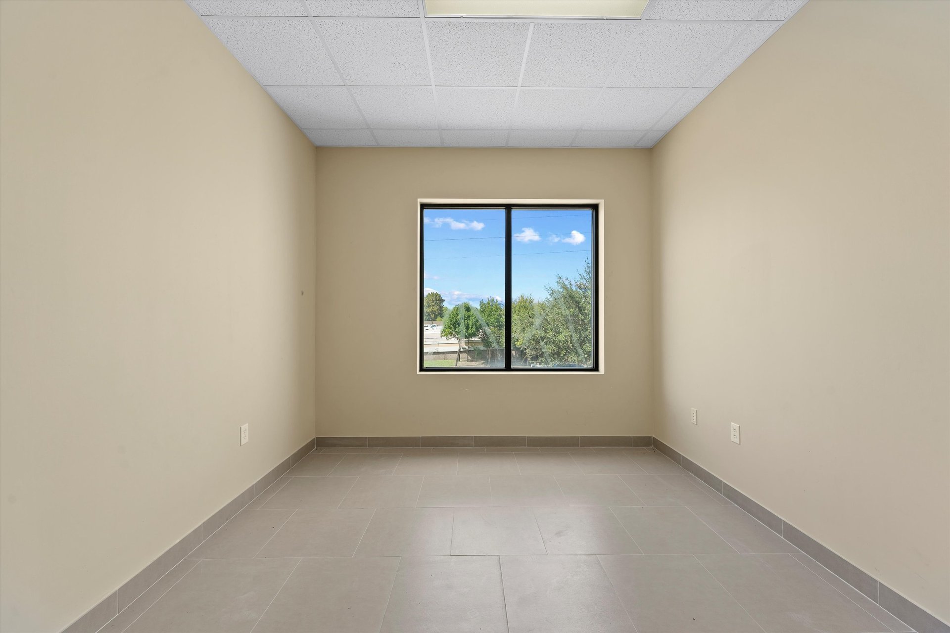 Vacant room with beige walls, gray tile flooring, and a black-framed window overlooking trees.