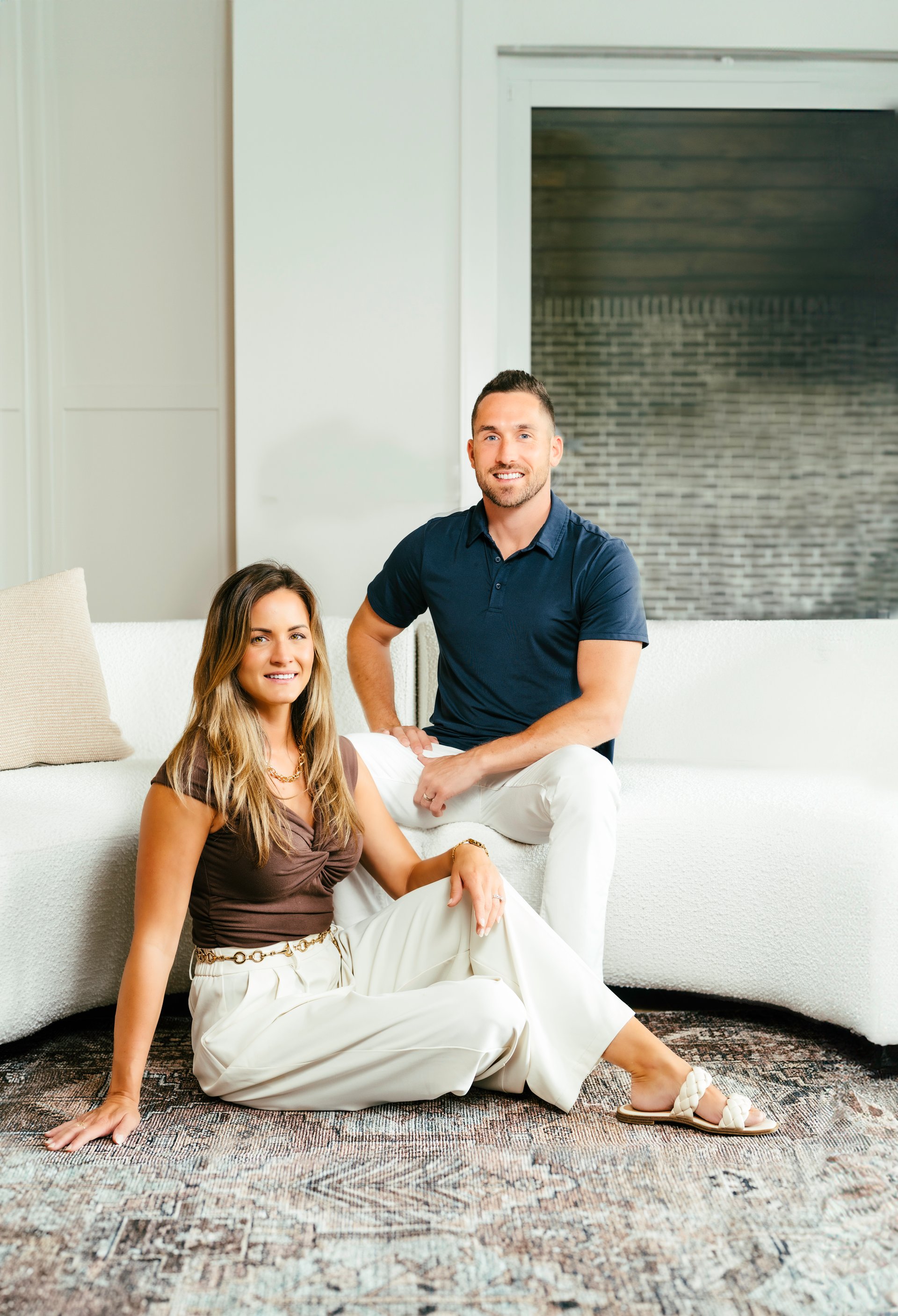 Two adults pose seated together in a bright living room.