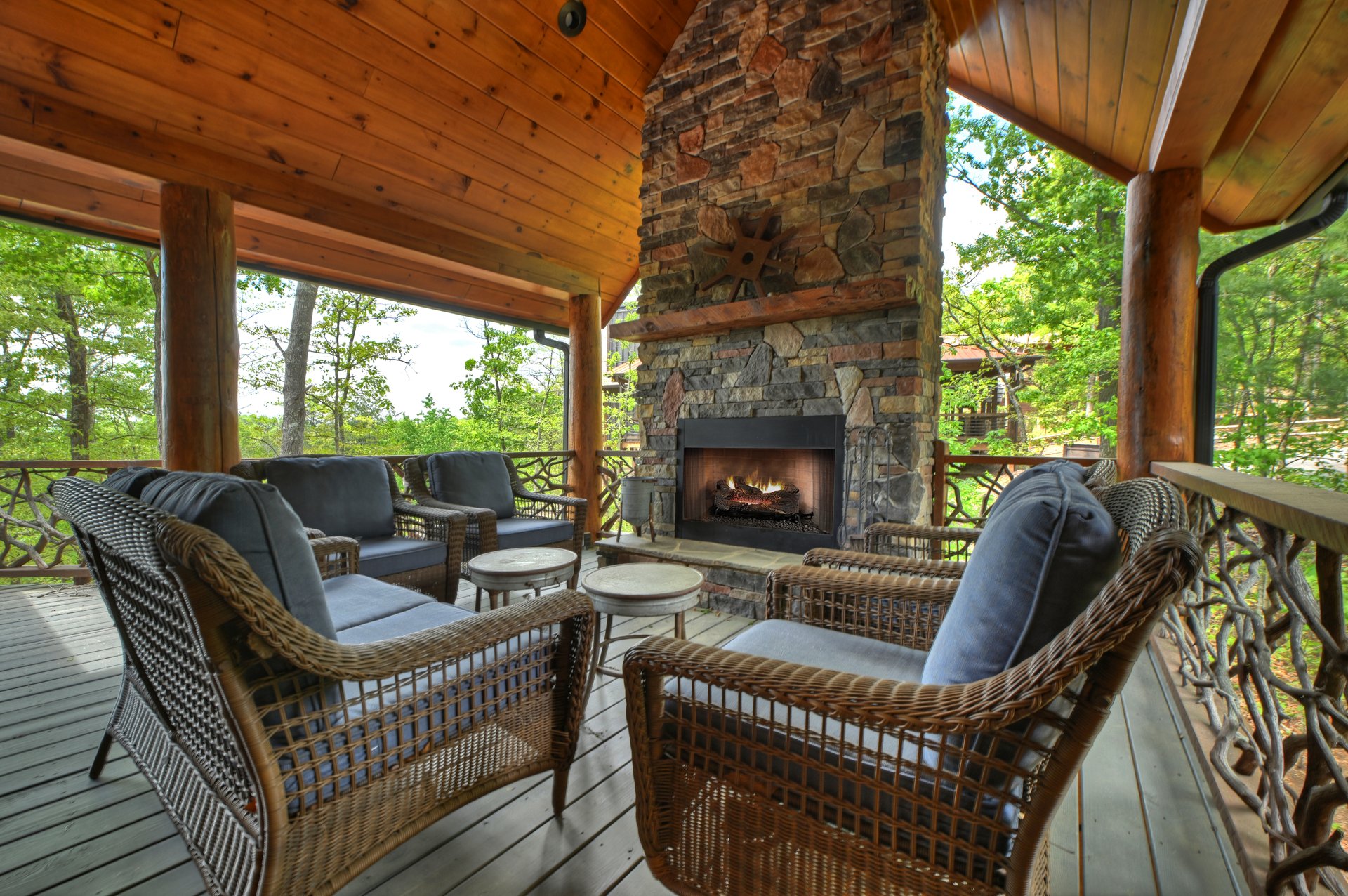 Covered deck with wicker seating around a stone fireplace, overlooking leafy woods.
