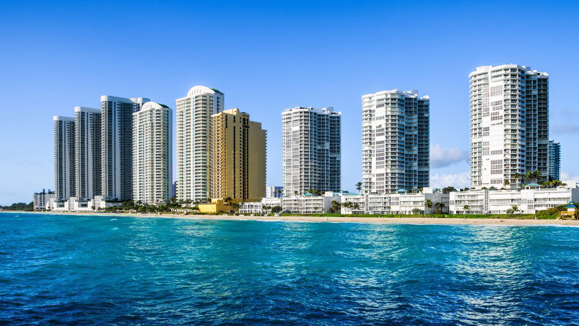 Panoramic view of white high-rise condominium buildings along Miami beachfront with crystal blue ocean water in foreground under clear sky, showcasing premium waterfront real estate listings by The Edit Real Estate