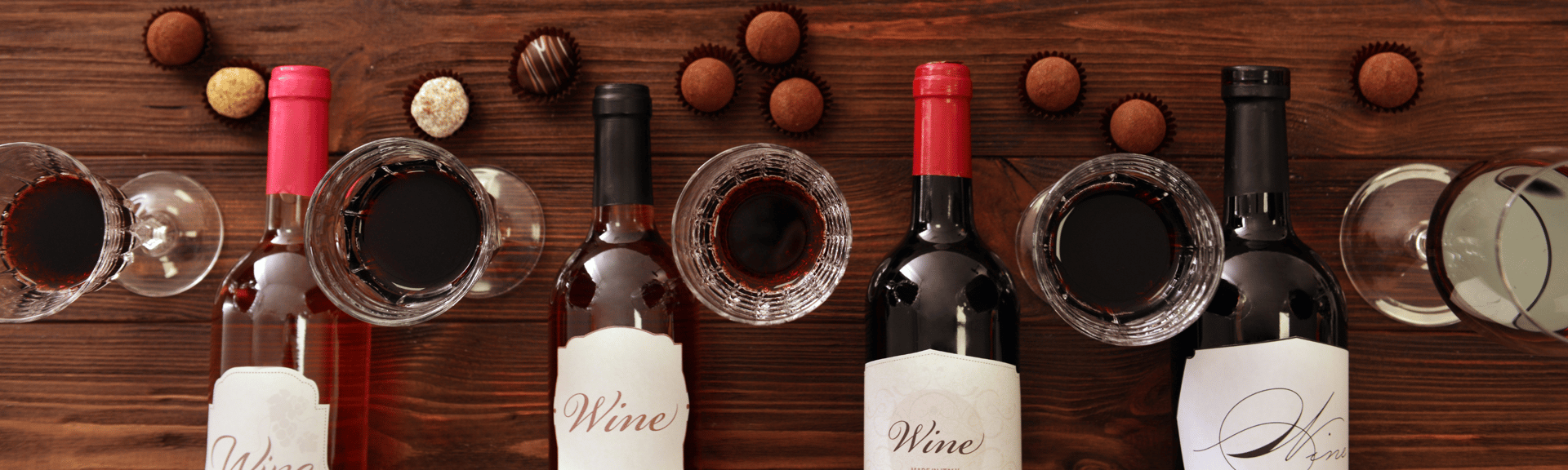 A wooden table displays several wine bottles, glasses filled with red wine, and assorted chocolates.