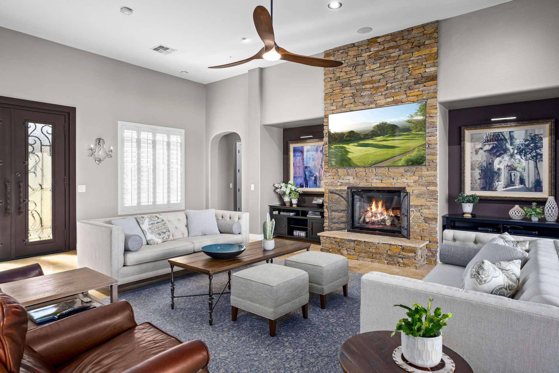 Staged living room with a stone fireplace, sofas, ottomans, and a large ceiling fan.