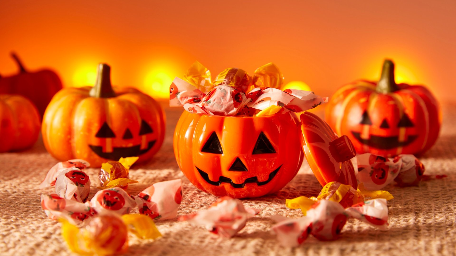 Orange jack-o-lantern Halloween pumpkin bucket filled with wrapped candy surrounded by glowing carved pumpkins with warm lighting