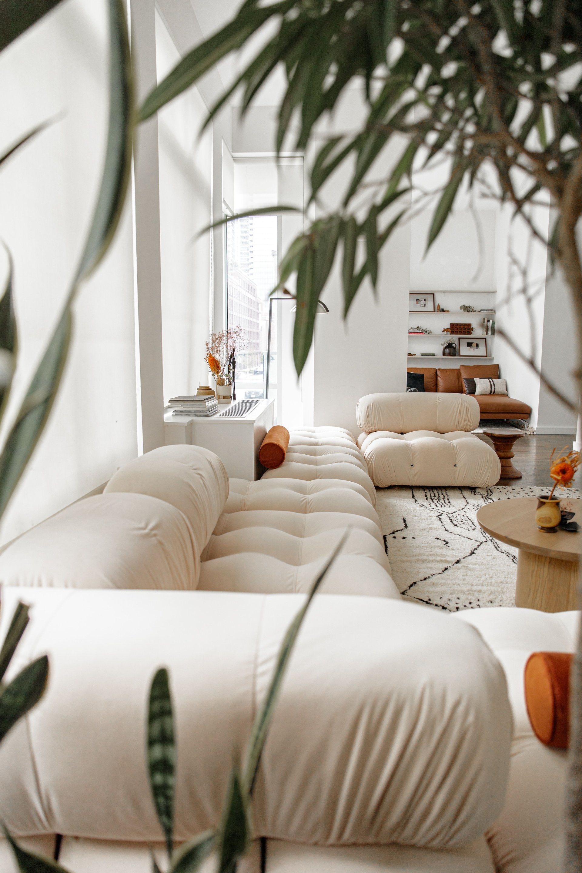 Bright living room with cream tufted sofas, rust-colored seating, built-in shelves, and leafy plants.