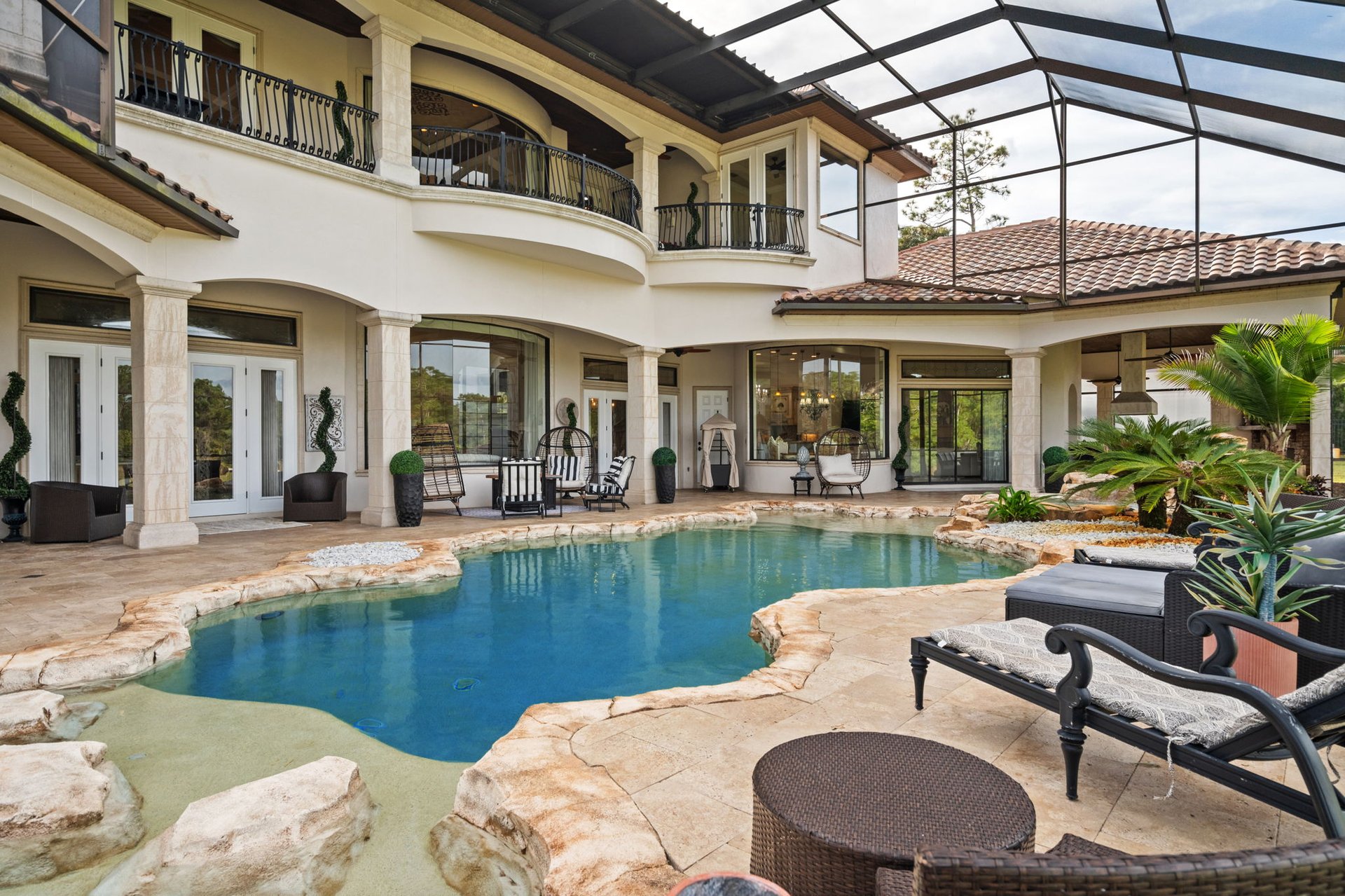 Freeform backyard pool and furnished patio beneath a screened enclosure beside a two-story home.