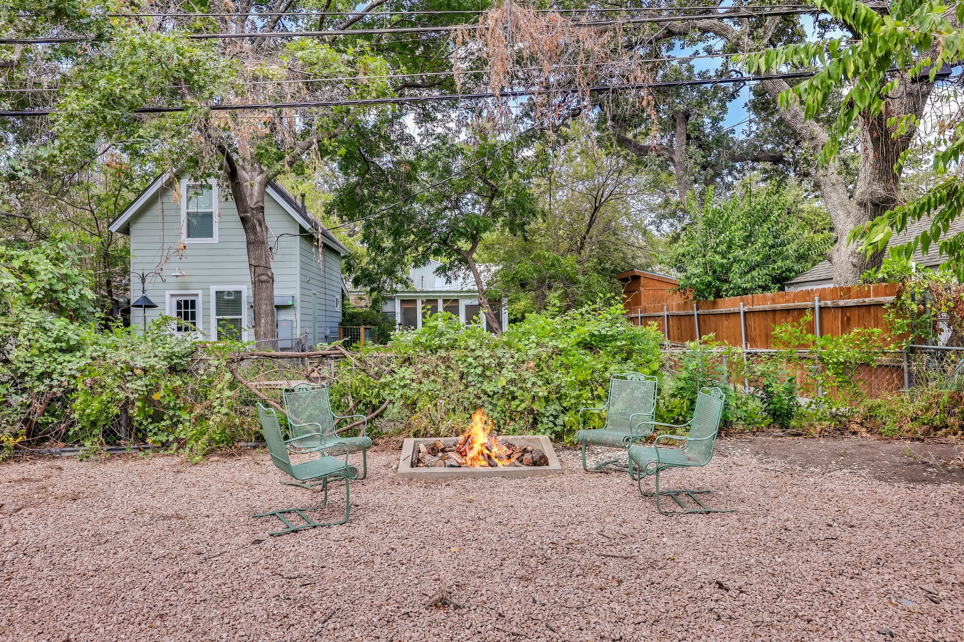 Gravel backyard with a lit fire pit, green chairs, and mature trees.
