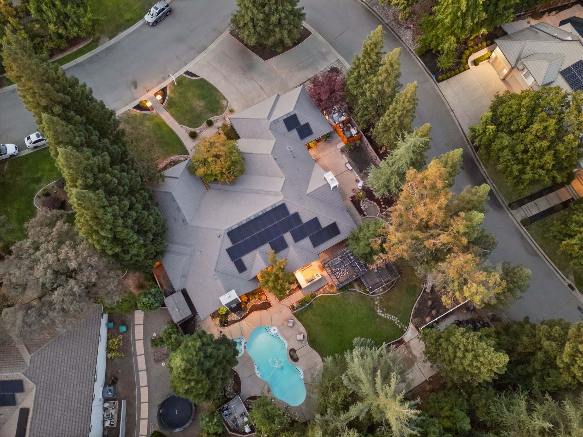 Aerial view of a home with rooftop solar panels, a backyard pool, patios, and mature trees.
