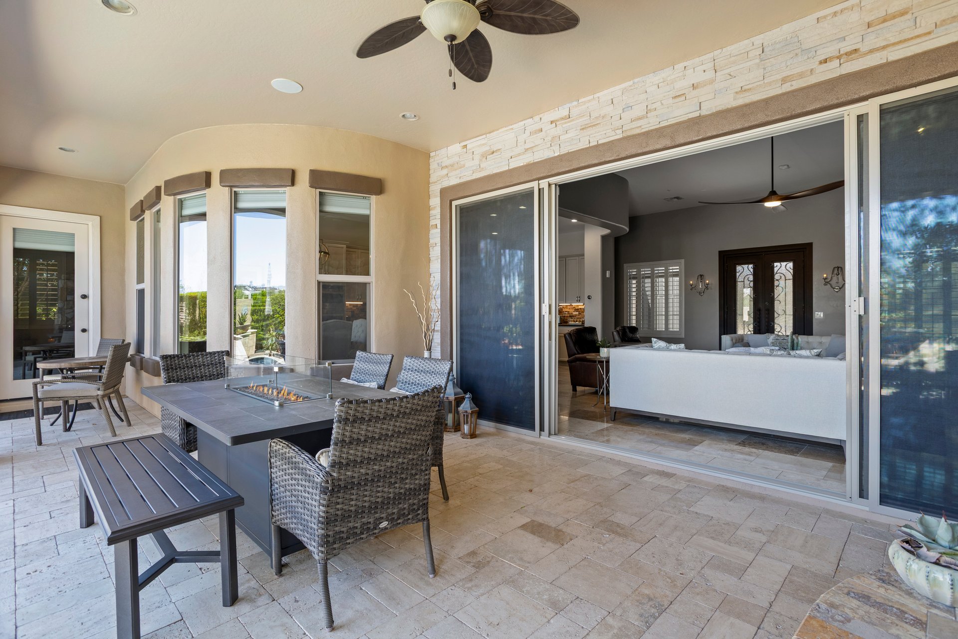 Covered outdoor patio with wicker seating, a fire table, stone tile flooring, ceiling fan, and large sliding doors opening to a living room.