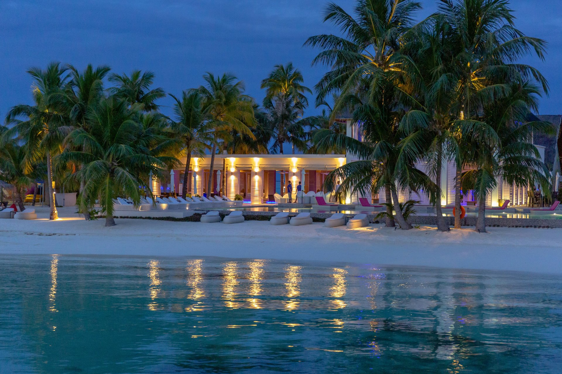 Illuminated beachfront resort with palm trees, lounge chairs, and reflections on the water at dusk.