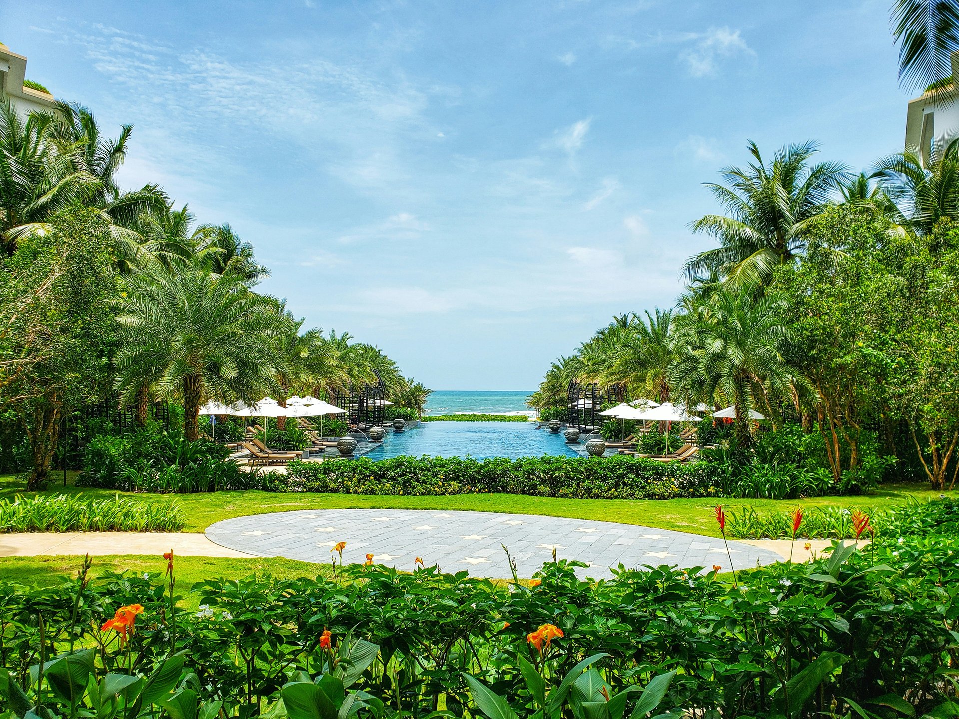 Symmetrical view of a tropical resort pool surrounded by palm trees, leading to the ocean in the distance.