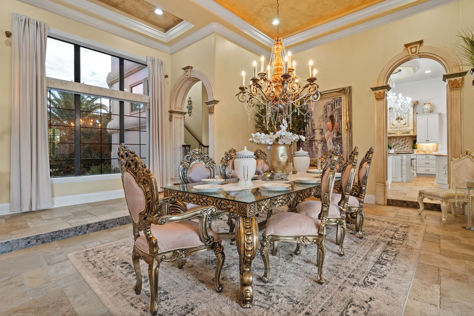 Furnished dining room with an ornate glass-top table, carved chairs, chandelier, and arched kitchen entrance.