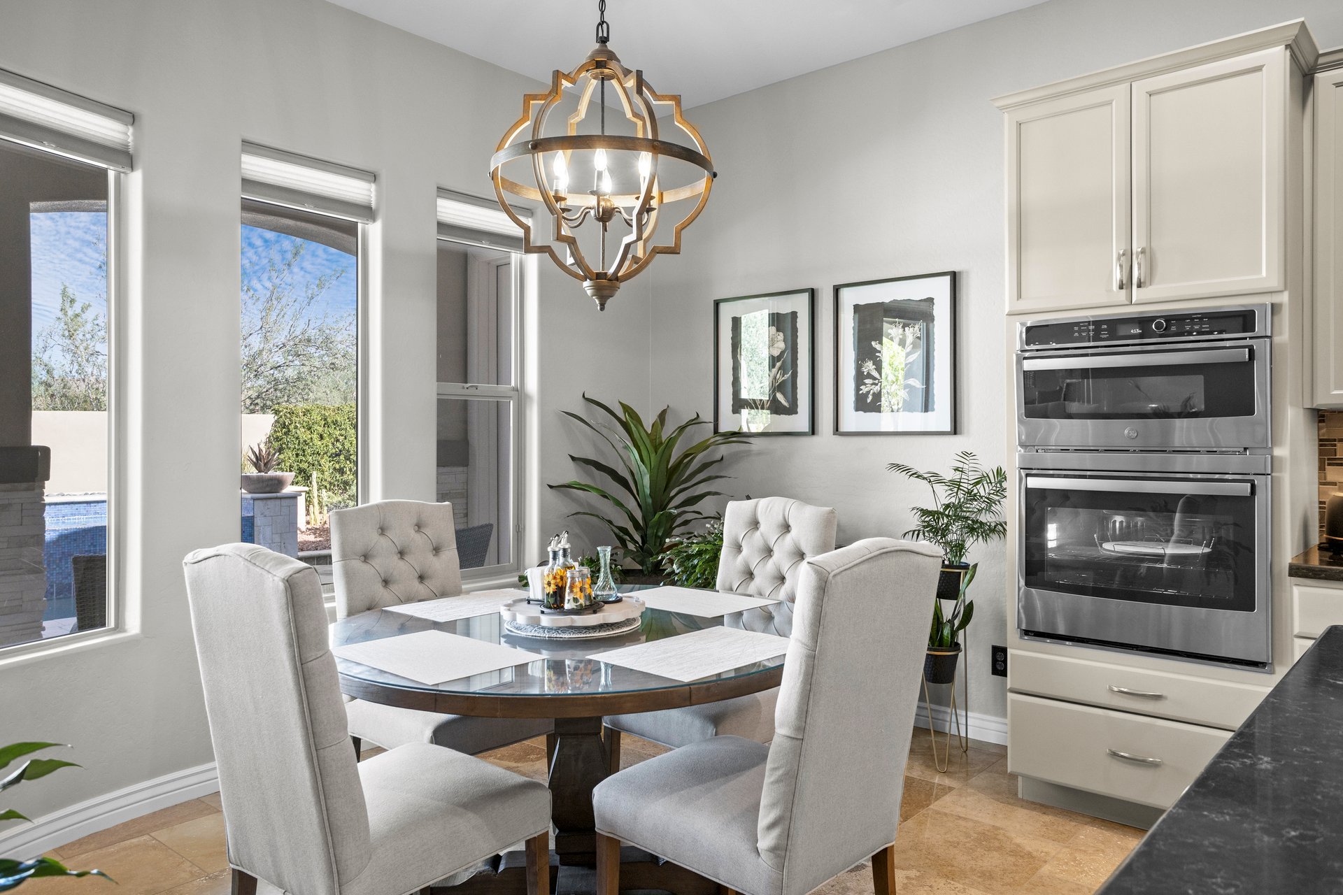 Bright dining area with a round glass table, four upholstered chairs, large windows, a decorative chandelier, and built-in double ovens beside the kitchen.