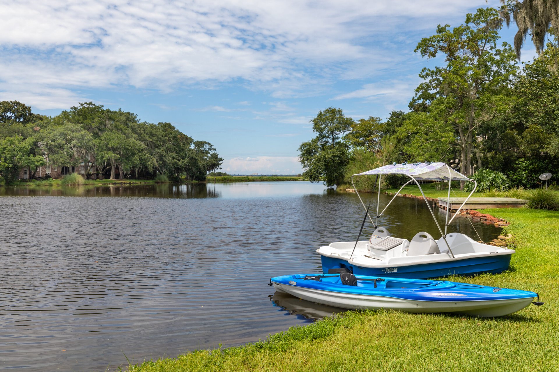 Tree-lined waterfront with a pedal boat and kayak beside the grassy shore.
