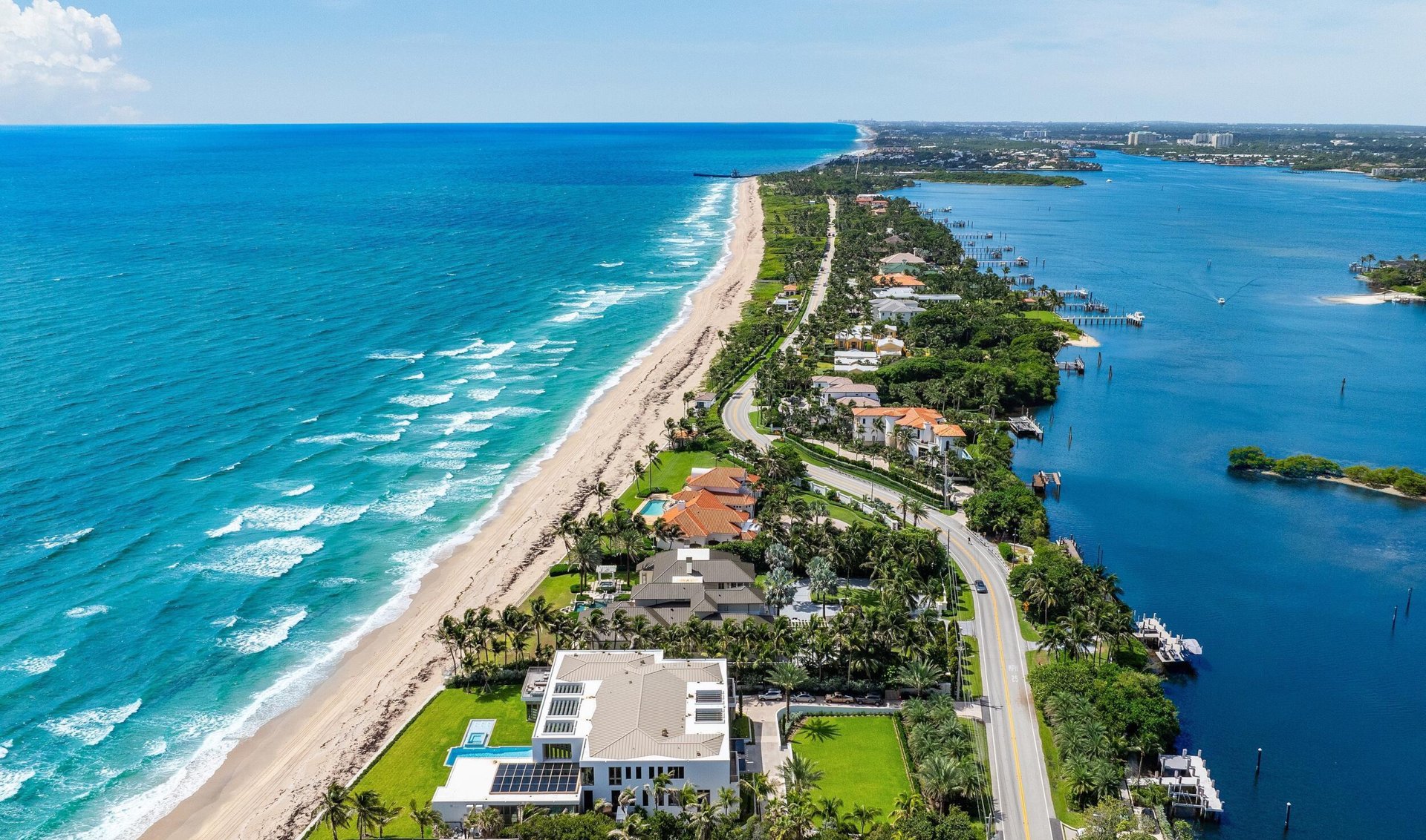 Aerial view of waterfront homes between an ocean beach and the intracoastal, with a winding palm-lined road.