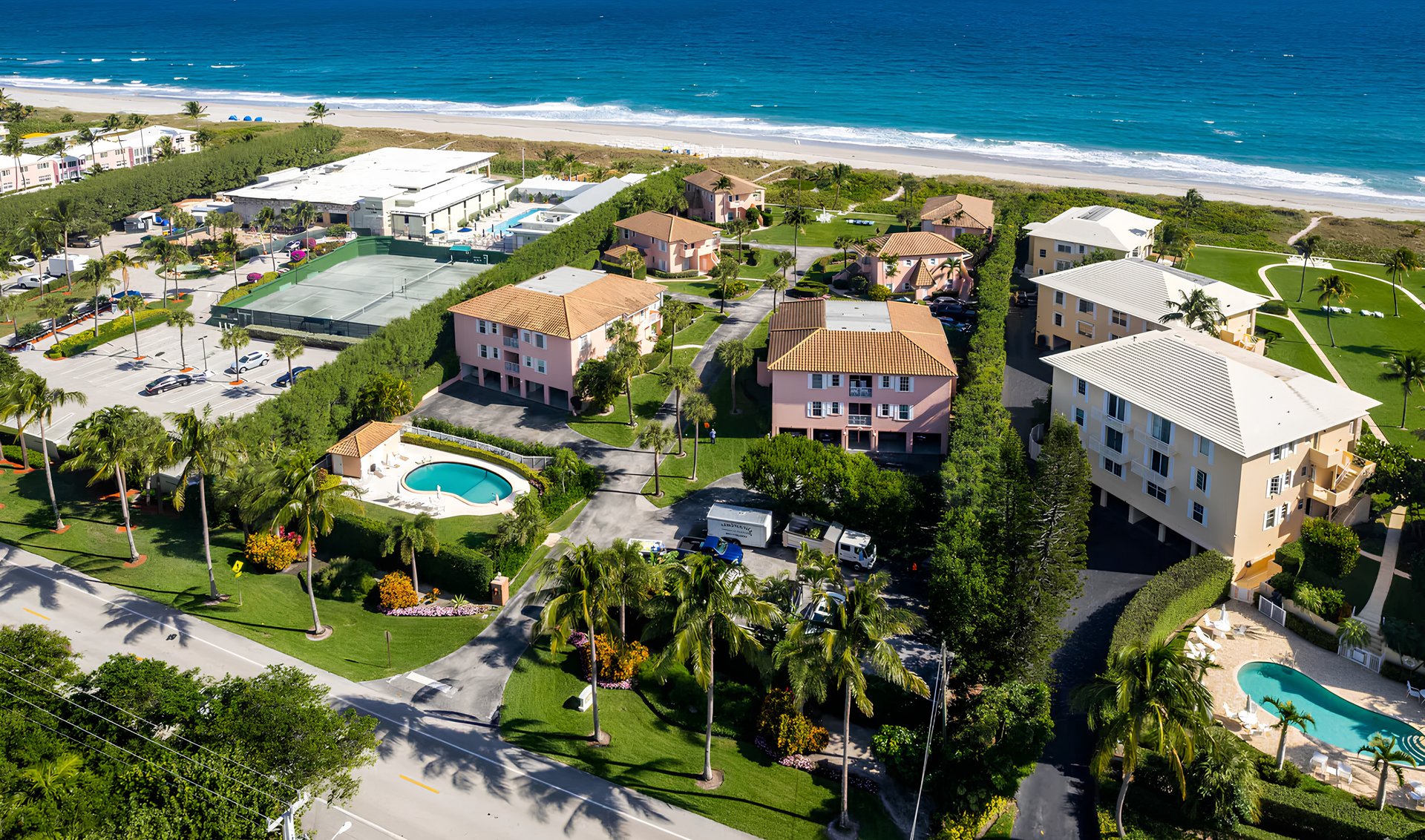 Aerial view of low-rise condos, pools, and a tennis court beside the ocean.