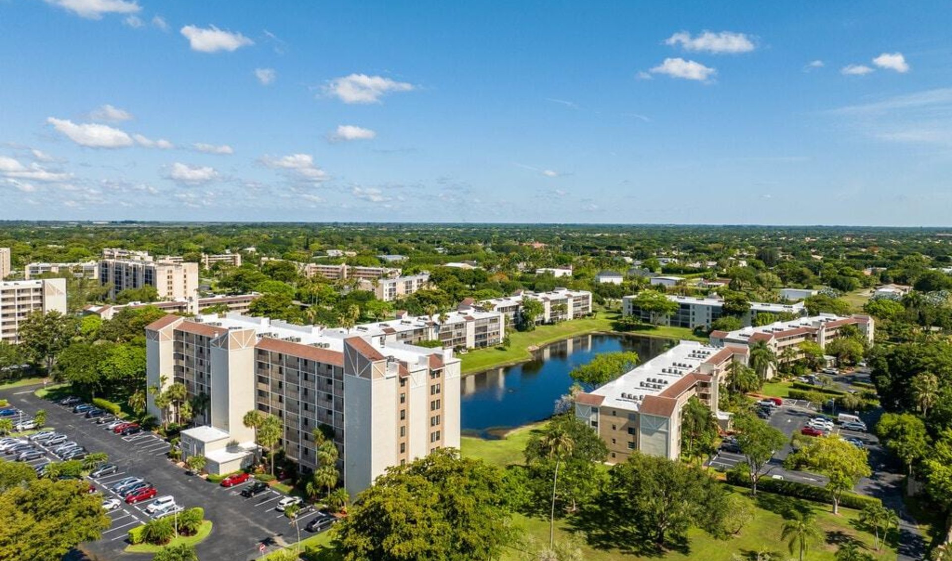 Aerial view of a condominium community surrounding a small lake, with landscaped grounds and parking lots.