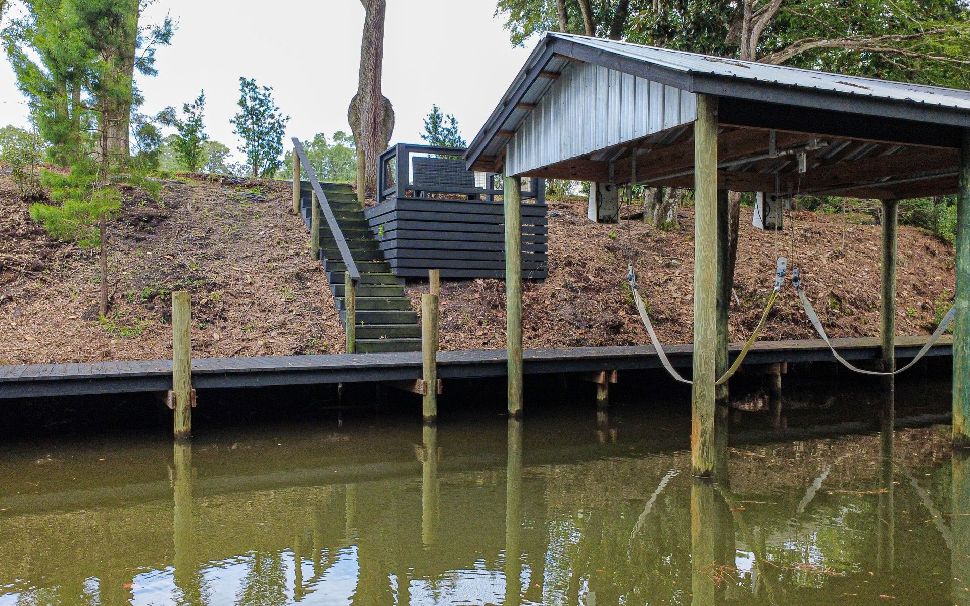 Covered boat lift and dock over calm water, with stairs climbing the wooded bank.