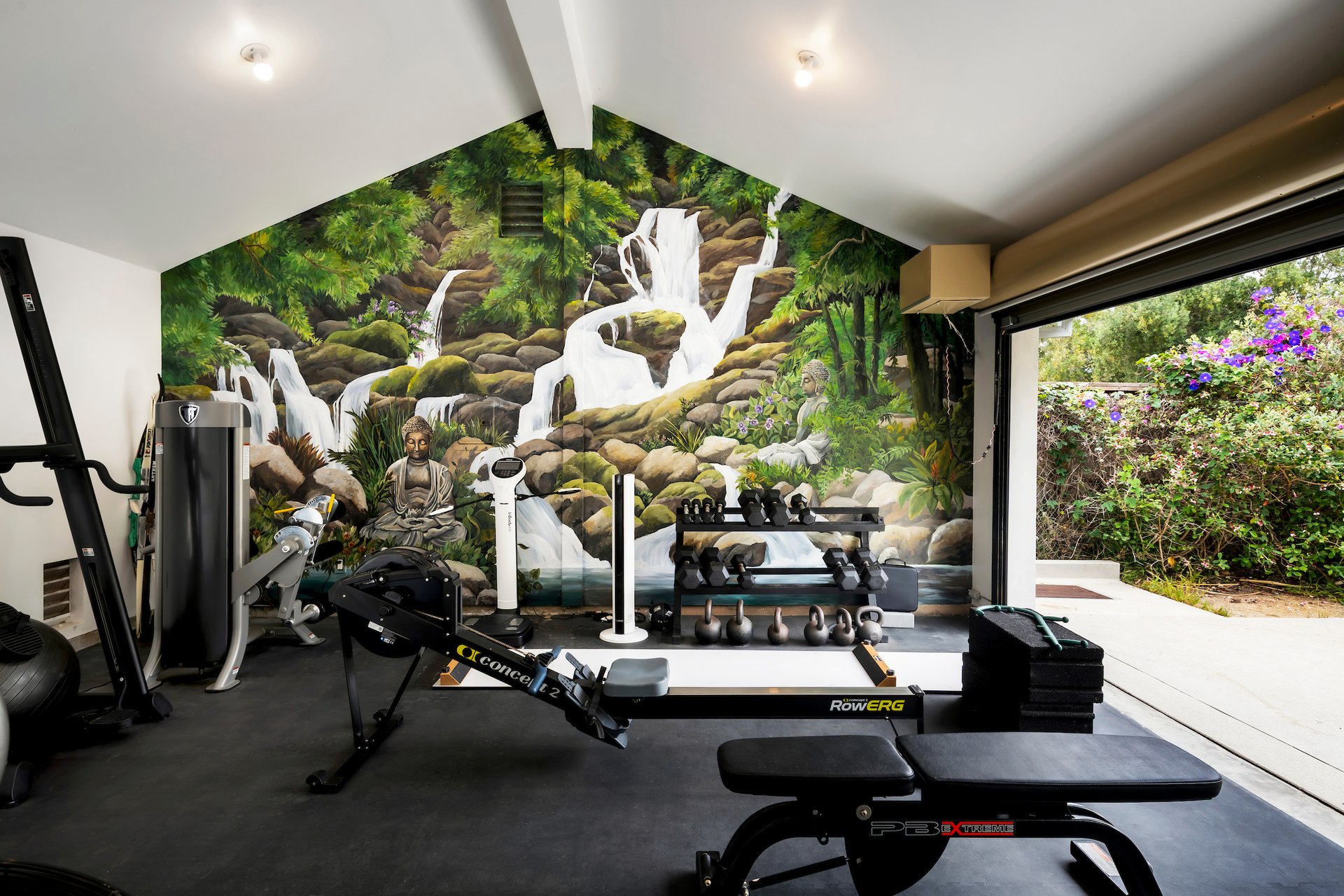 Home gym with rowing and weight equipment facing a forest waterfall mural and an open garden doorway.