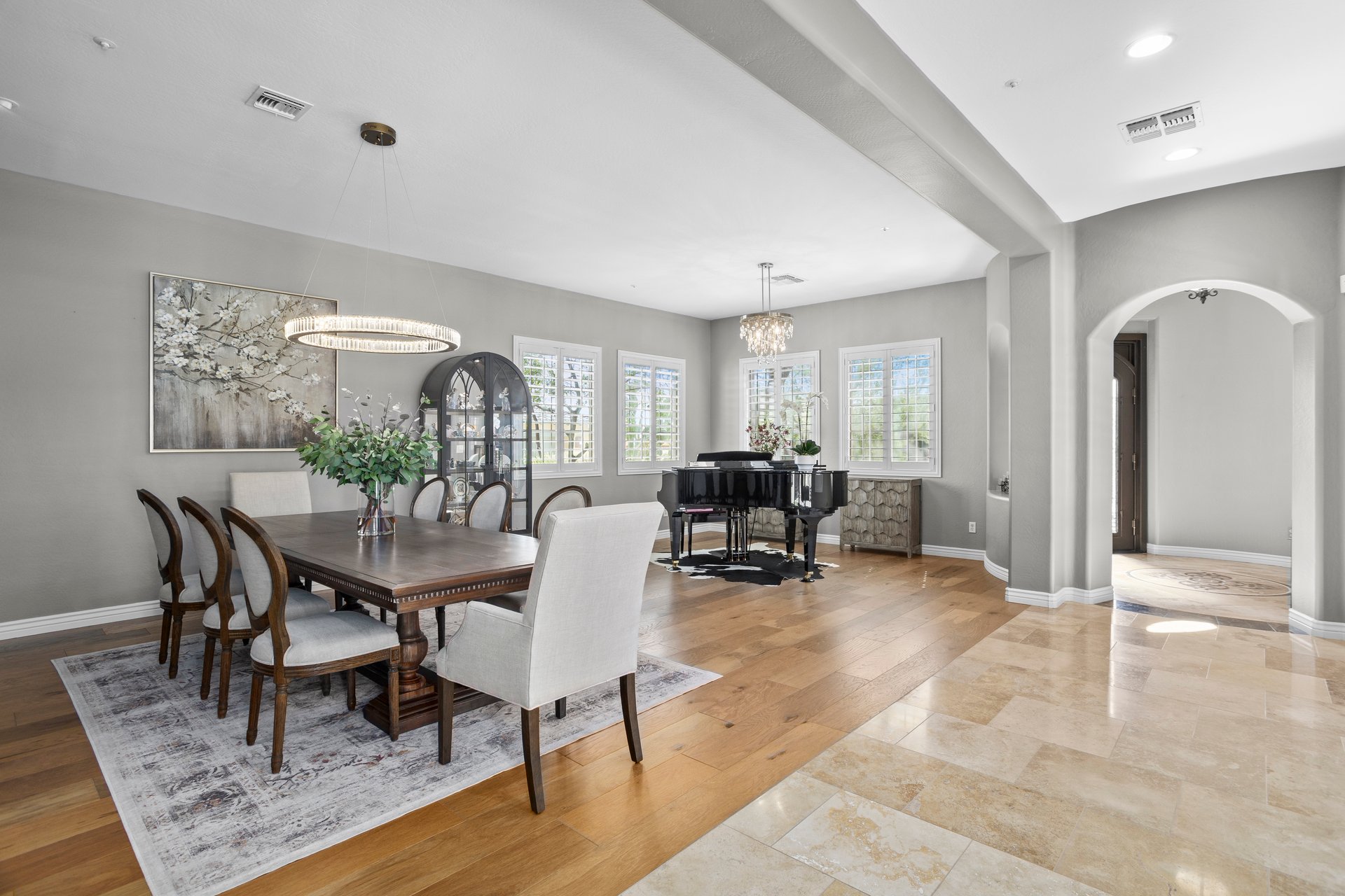 Bright formal dining room with a large table, upholstered chairs, chandelier, piano, hardwood and tile flooring, and arched openings to a foyer area.