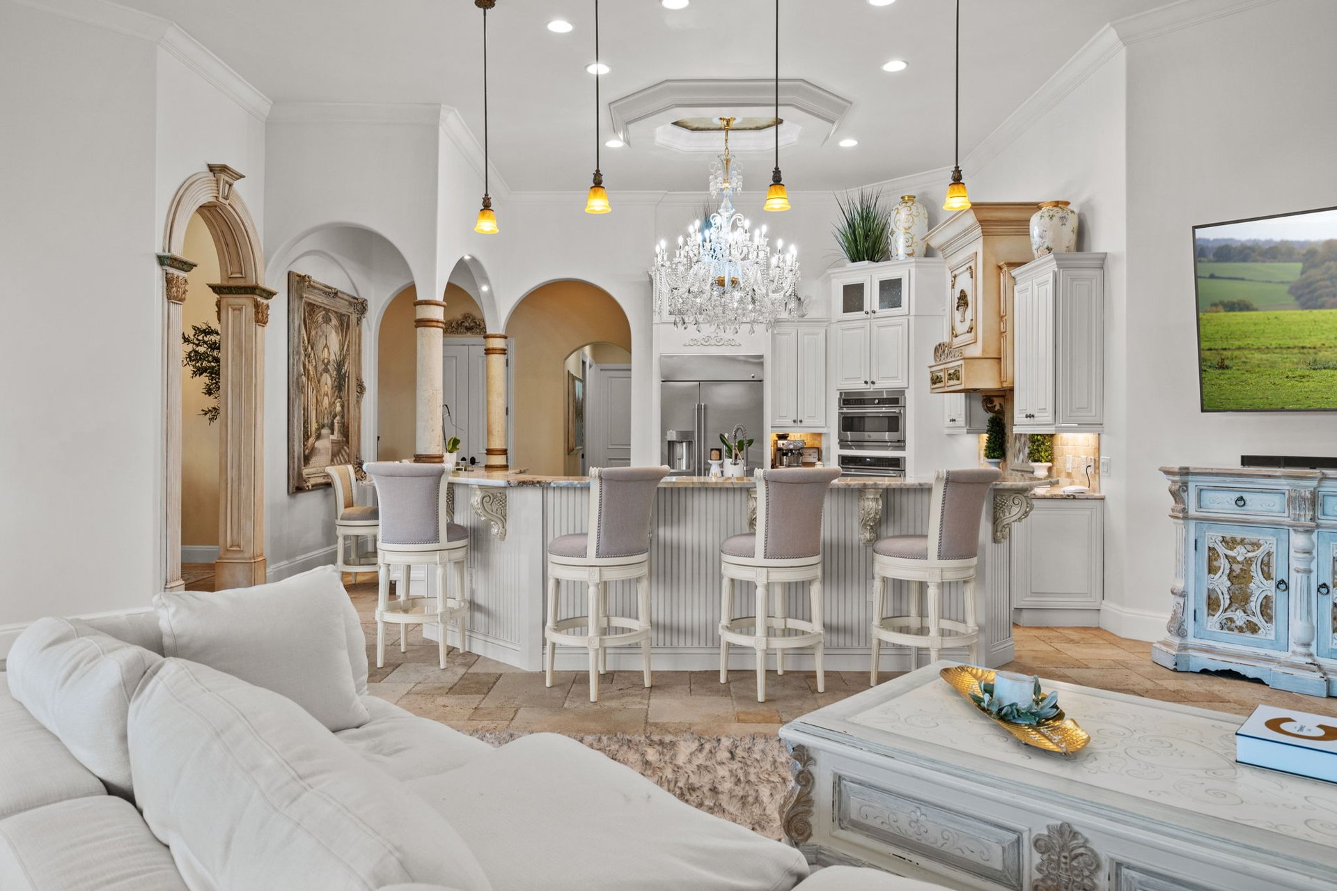 Ornate kitchen with a long island, four stools, chandelier, and adjoining white living room.