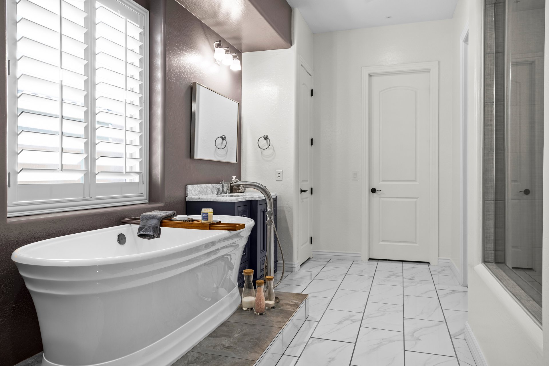 A bright modern bathroom with a freestanding tub beside a vanity, white tile floors, a glass shower, and plantation shutters on the window.