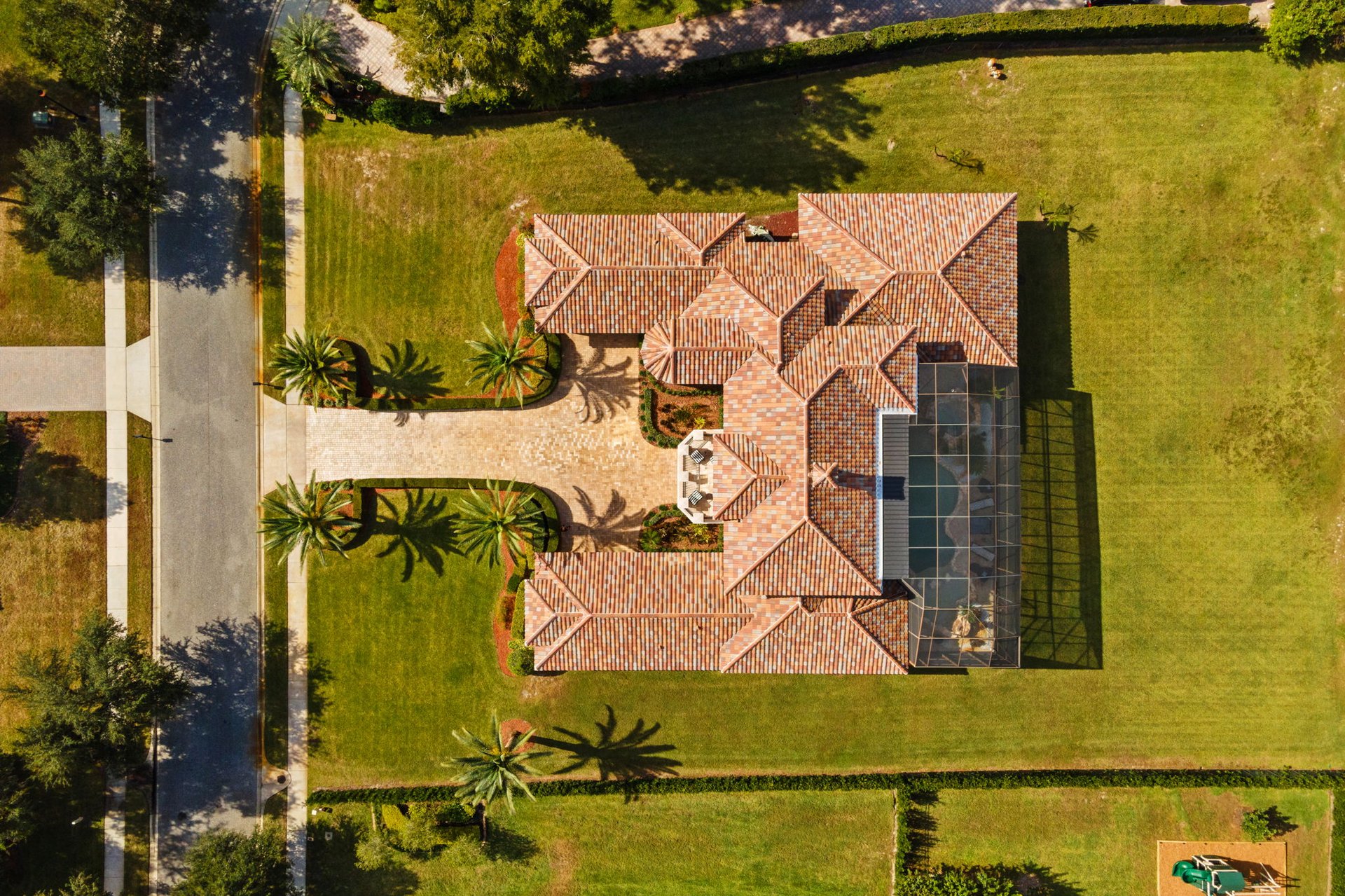 Aerial view of a tile-roofed home with a screened enclosure, palm-lined driveway, and expansive lawn.