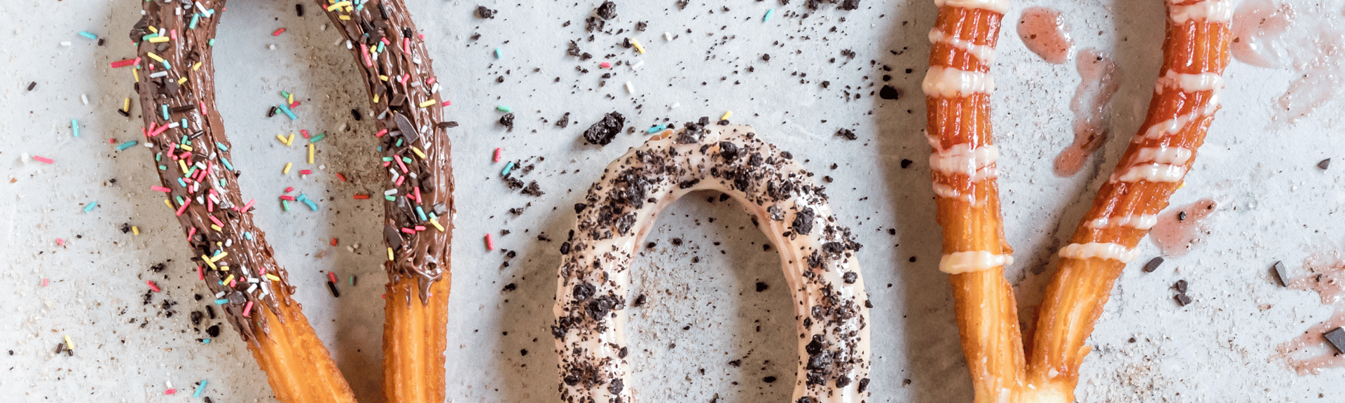 Three decorated churros on a light surface: one with chocolate and sprinkles, one with cream and crushed cookies, and one with white icing.