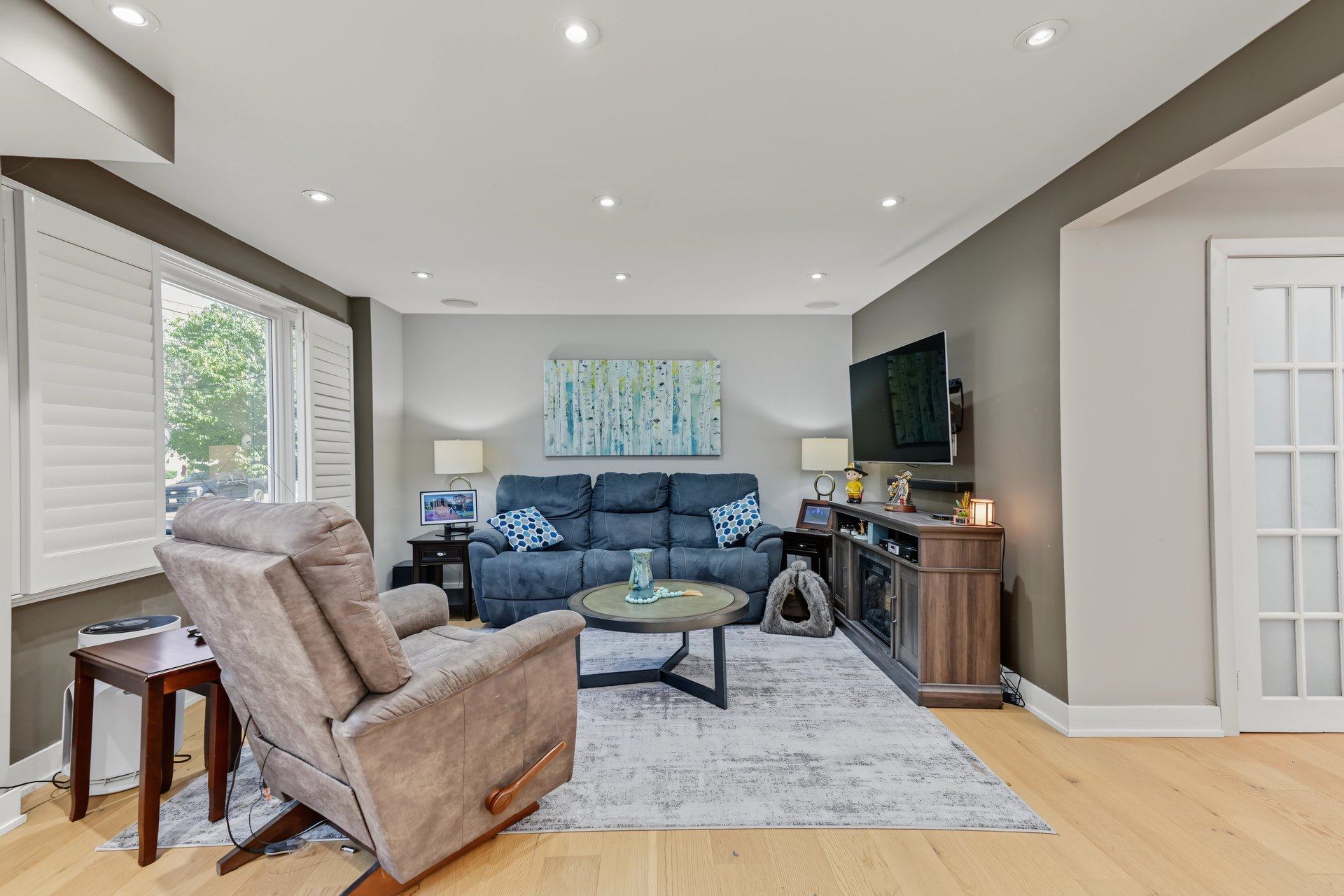 Living room with a blue sofa, leather recliner, wall-mounted television, and large shuttered window.