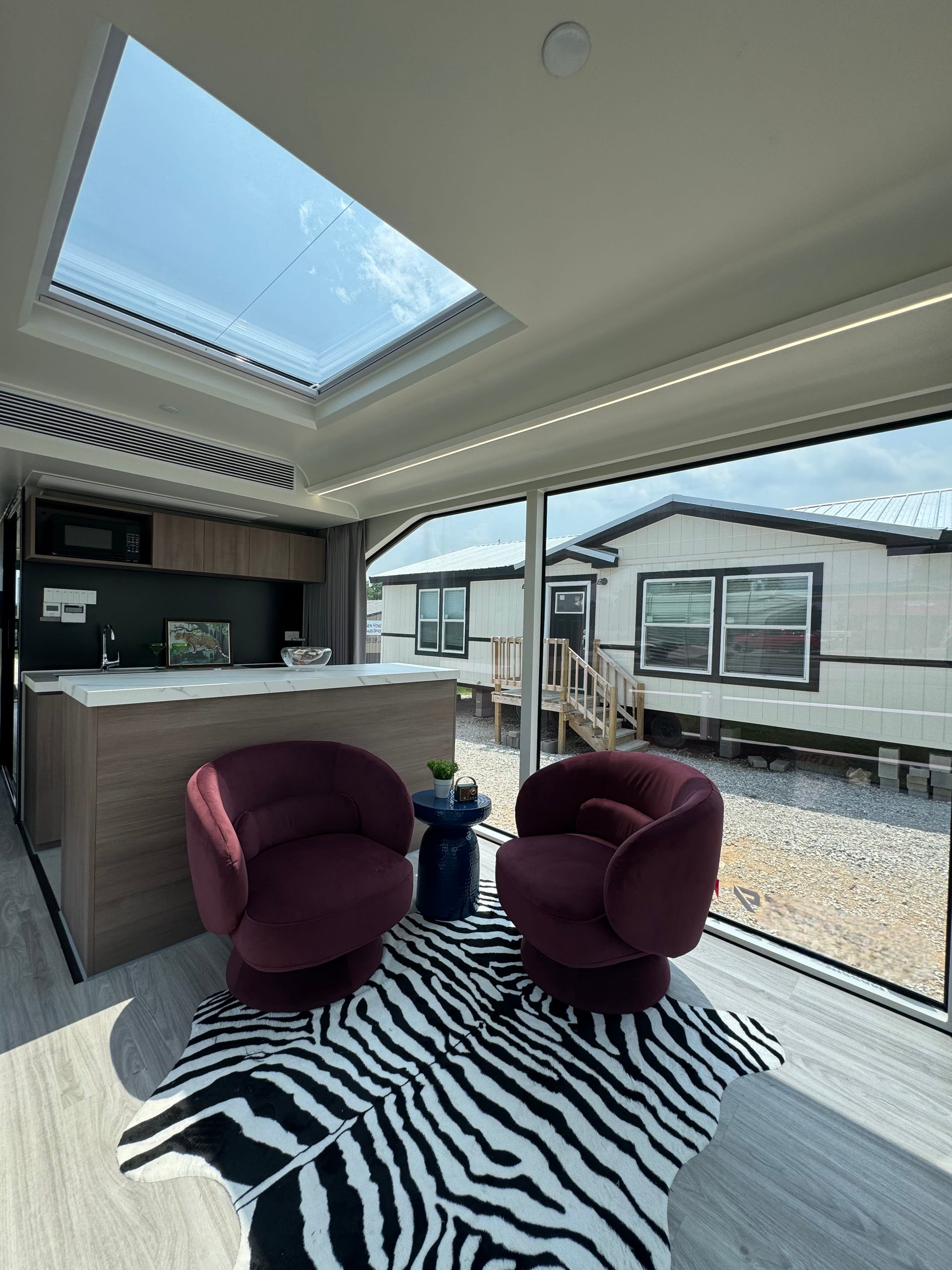 Compact living area with burgundy chairs, a kitchenette, zebra-pattern rug, skylight, and large windows.