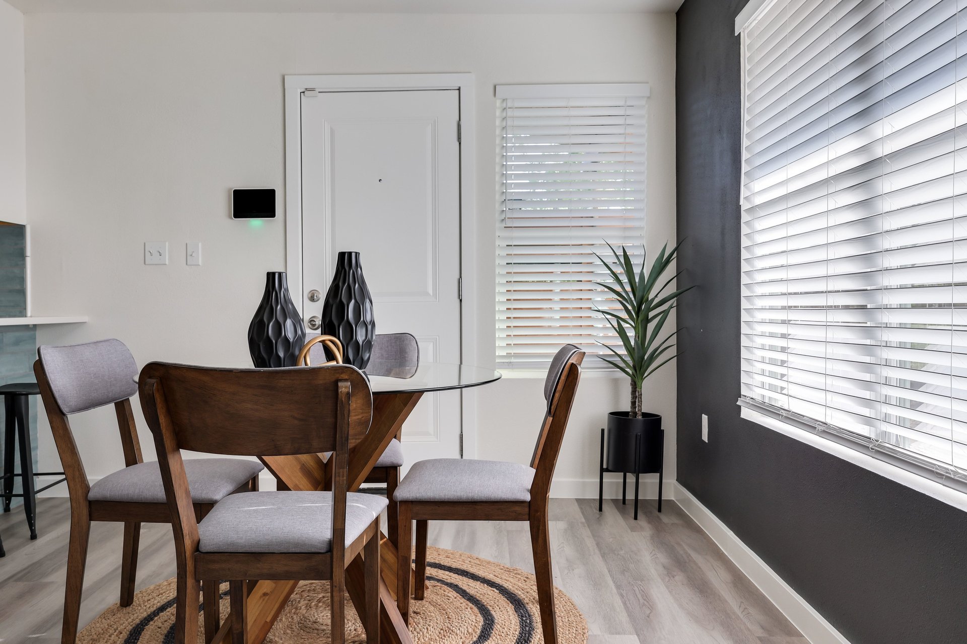 Dining area with a glass-top table, dark wood chairs, and a potted plant beside window blinds.