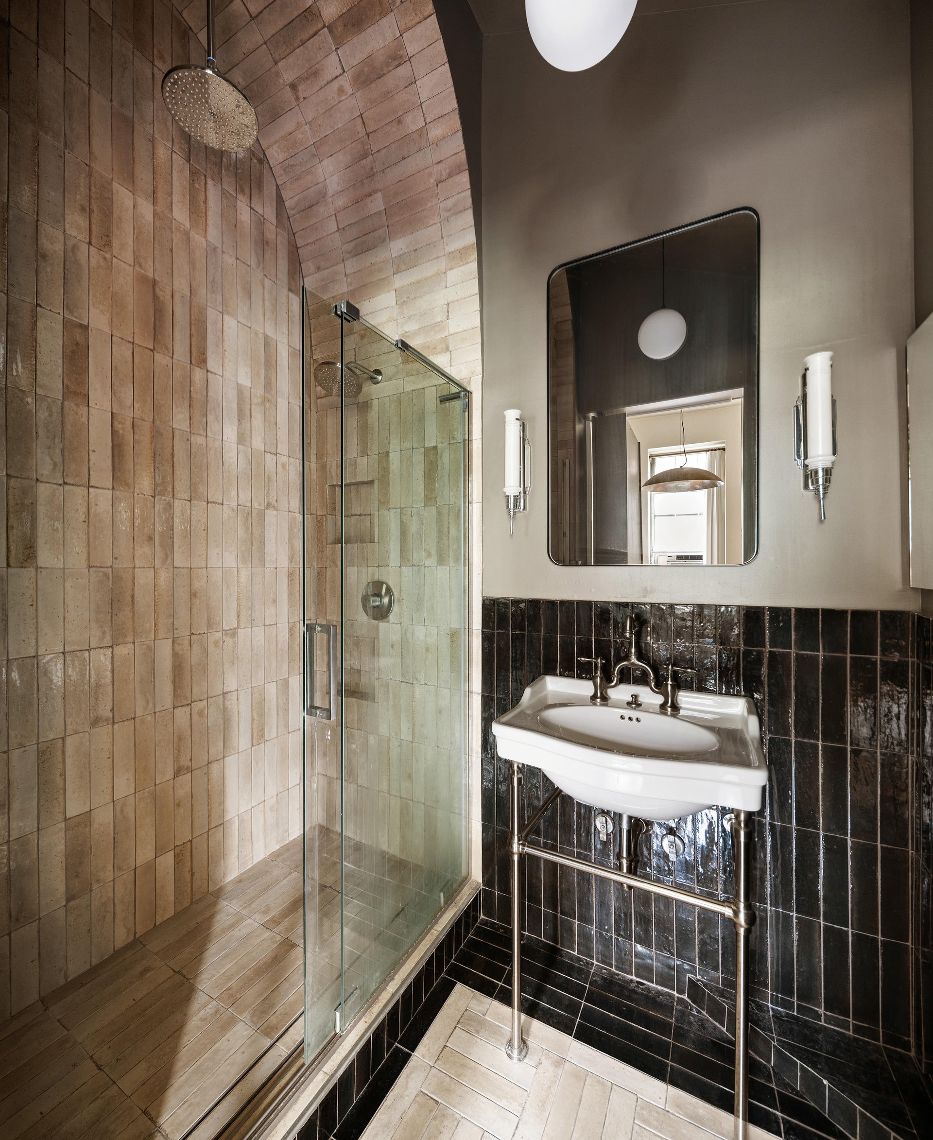 Bathroom with an arched tiled shower, glass enclosure, wall-mounted sink and dark tiled wainscot.