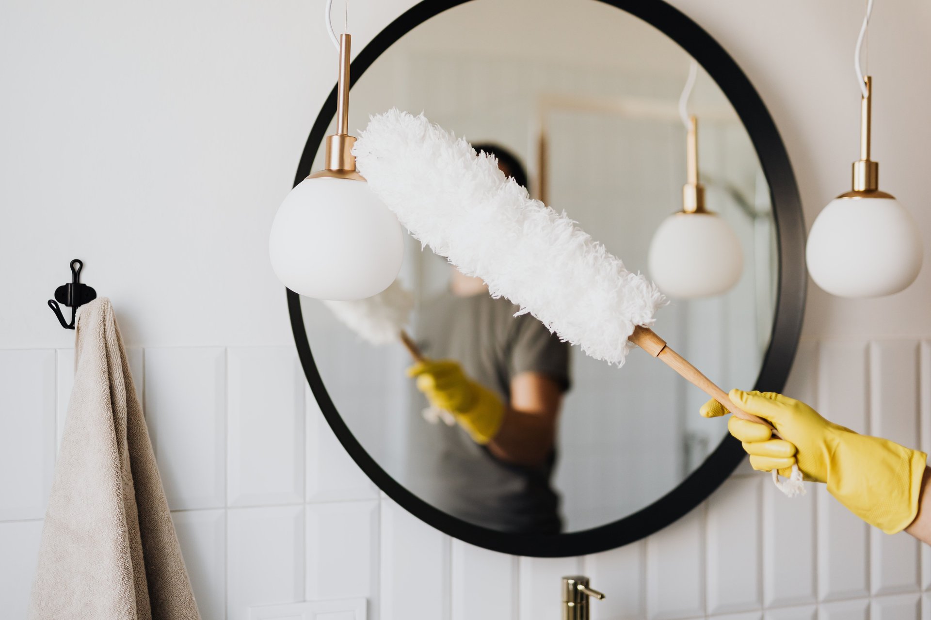 A gloved hand dusts a bathroom pendant light beside a round mirror.