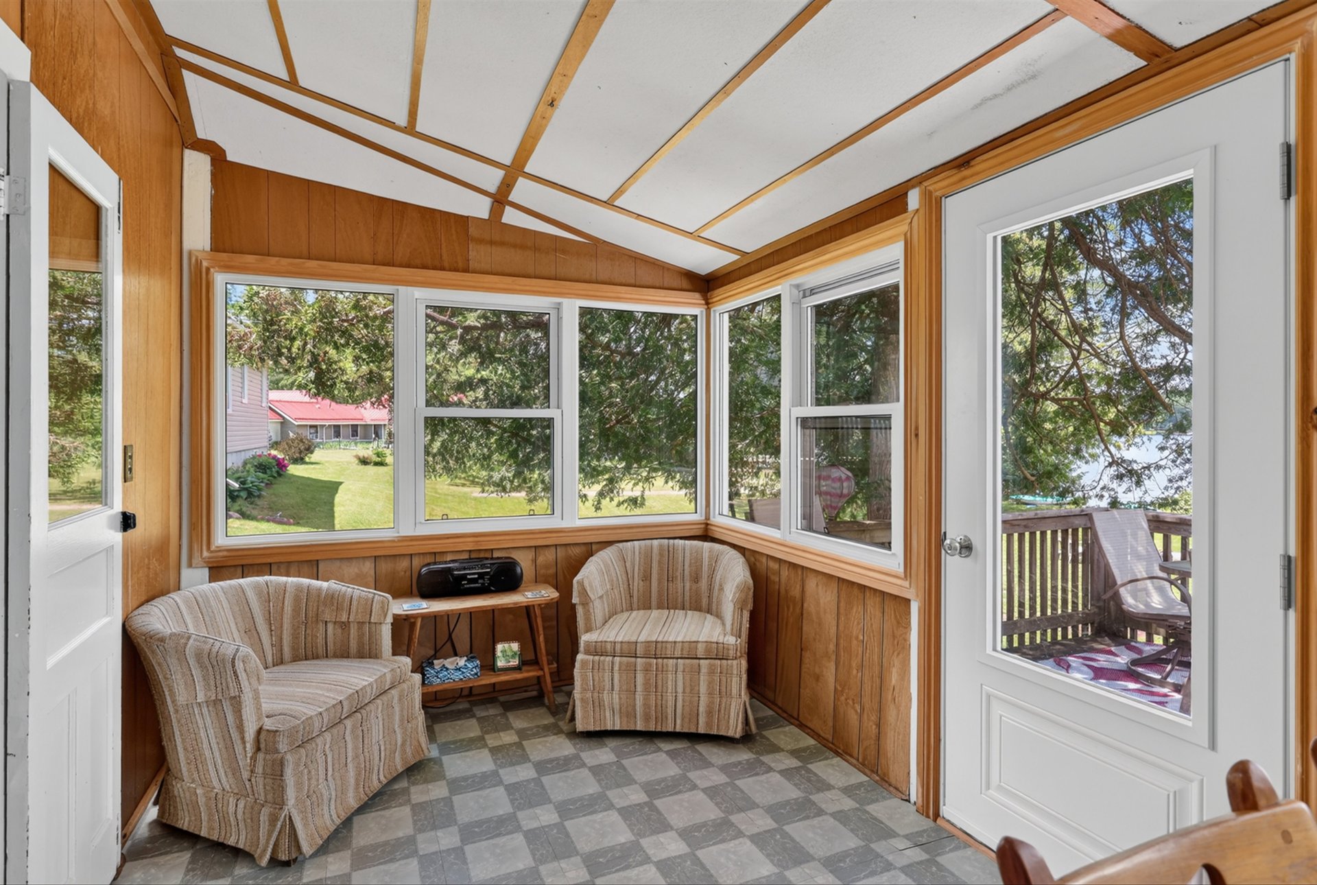 Sunroom with two upholstered armchairs, wood-paneled walls, and windows overlooking a lawn and deck.