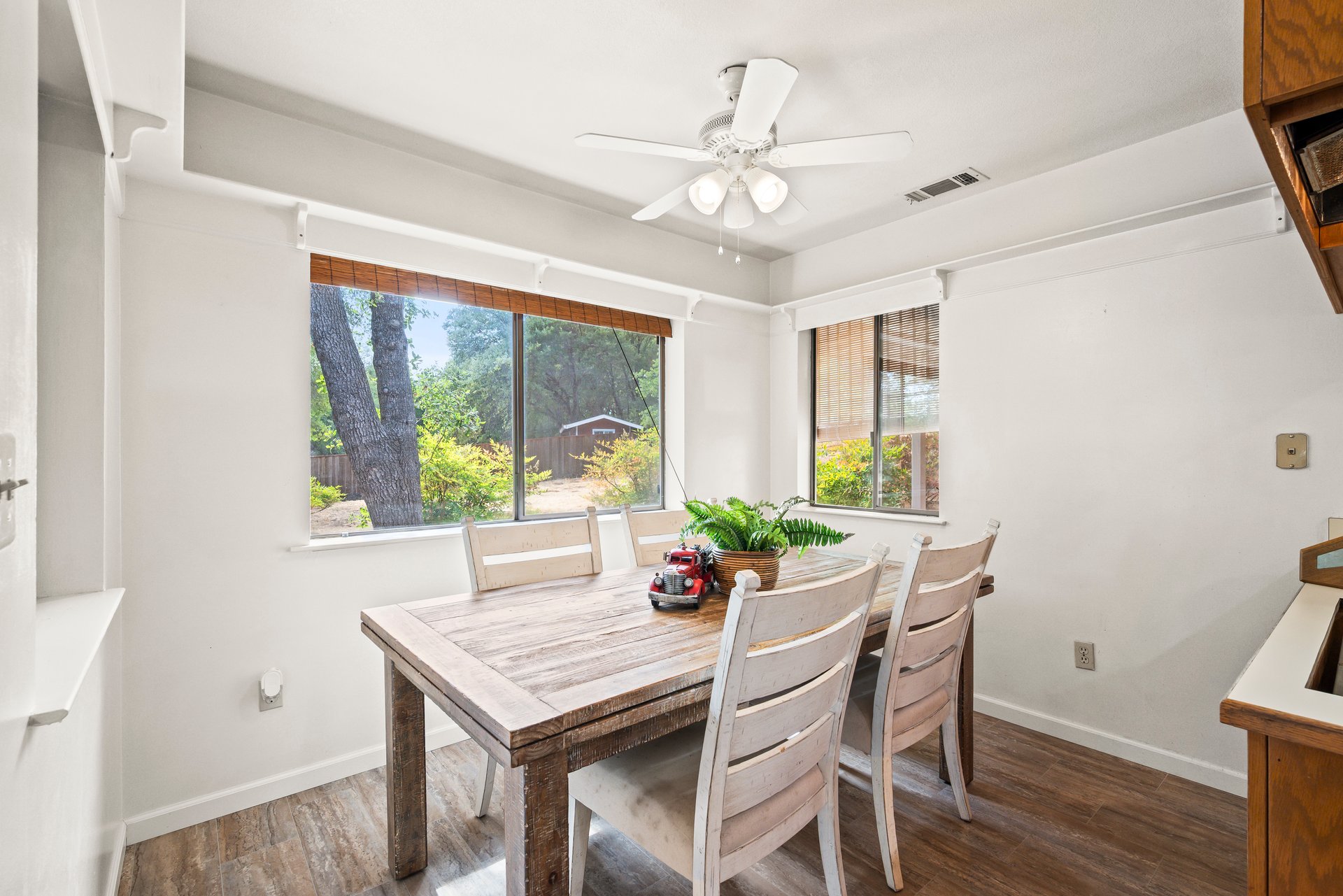 Dining area with a rustic wooden table, pale chairs, and windows overlooking a leafy yard.