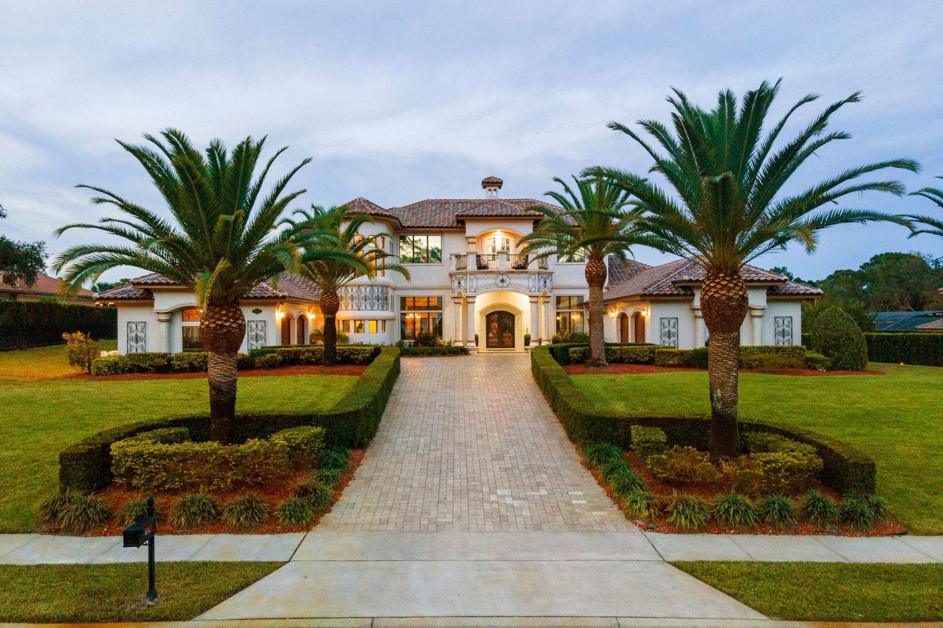 Mediterranean-style two-story home framed by palms, with a broad paved driveway and landscaped lawn.