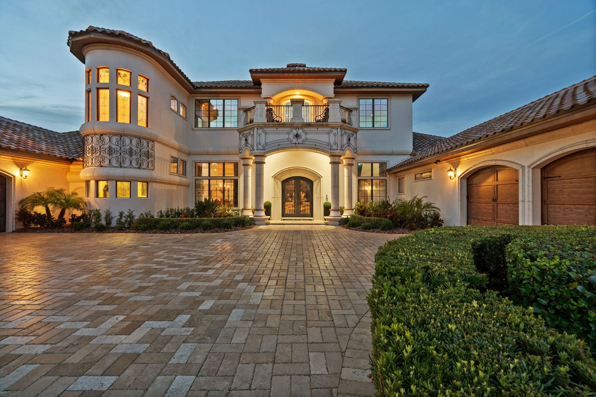 Two-story Mediterranean-style home with curved tower, ornate balconies, and broad paver driveway at dusk.