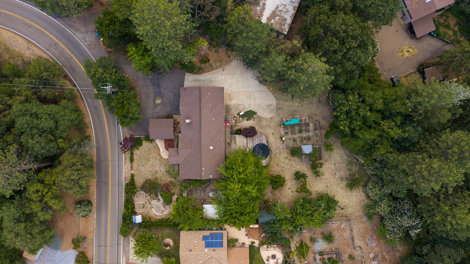 Aerial view of a wooded home beside a curving road, with a broad driveway and garden beds.