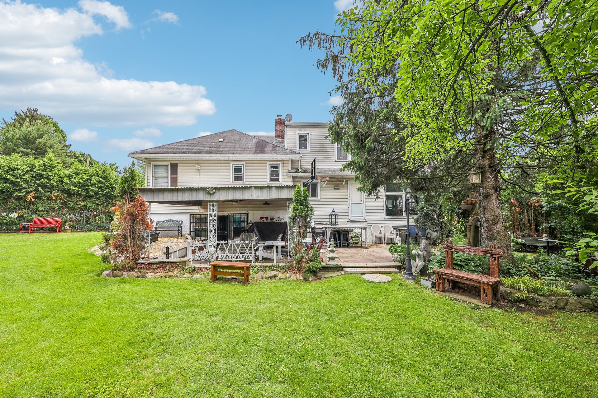 Rear of a two-story house with a covered patio, outdoor seating, and a broad lawn beneath mature trees.