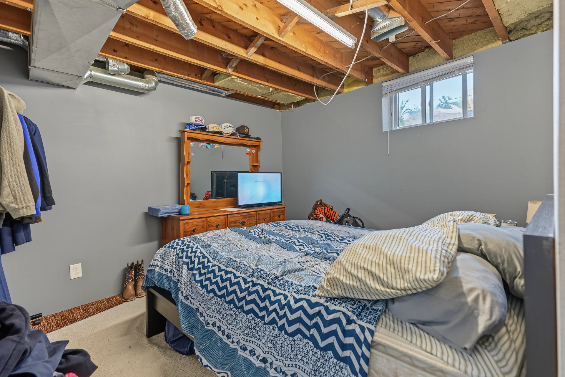 Bedroom with blue patterned bedding, wooden dresser, high window, and exposed ceiling joists.