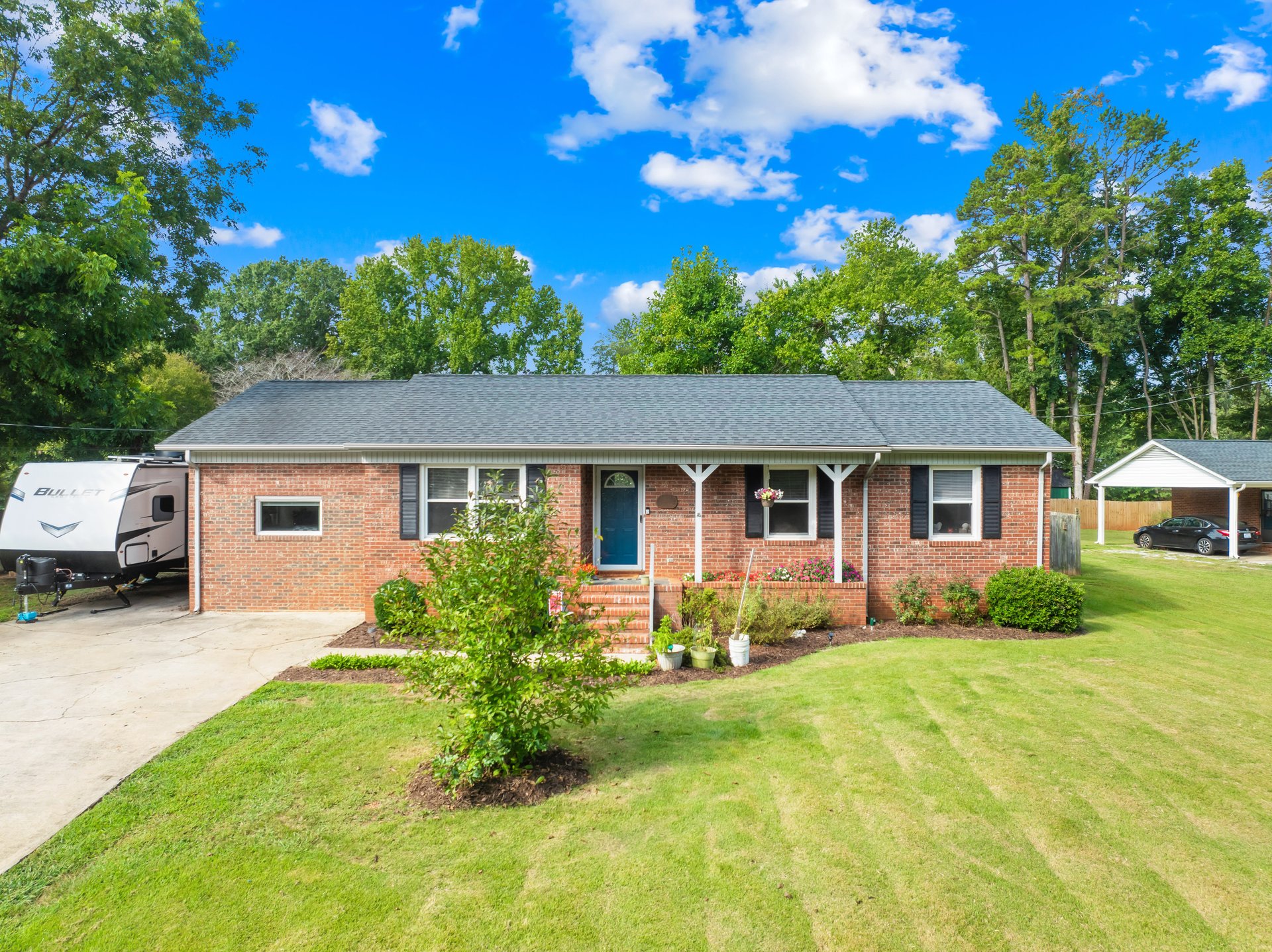 Brick ranch home with a blue front door, covered porch, broad lawn, and travel trailer in the driveway.