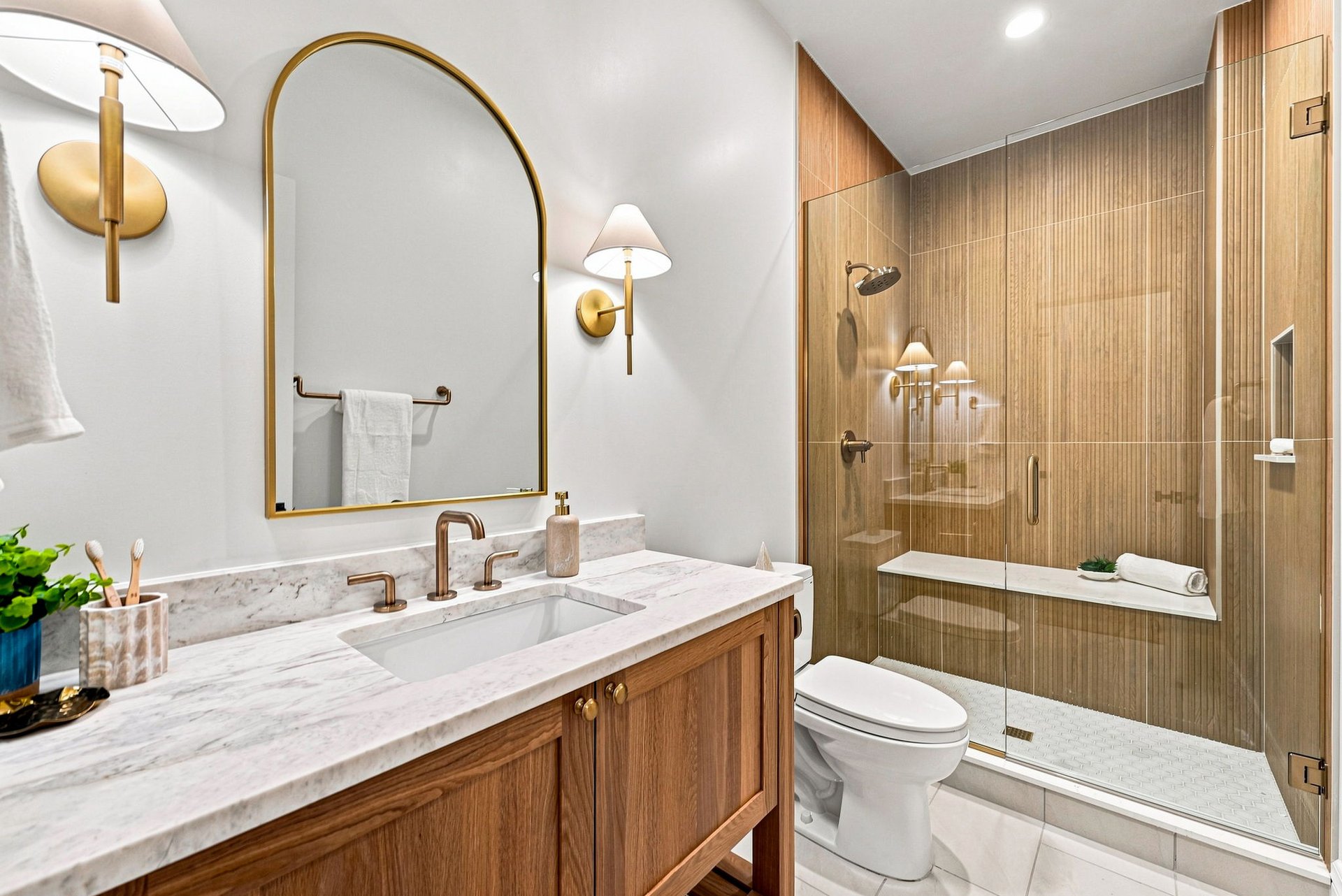 Bathroom with a marble vanity, brass fixtures, arched mirror, and wood-look tiled shower.