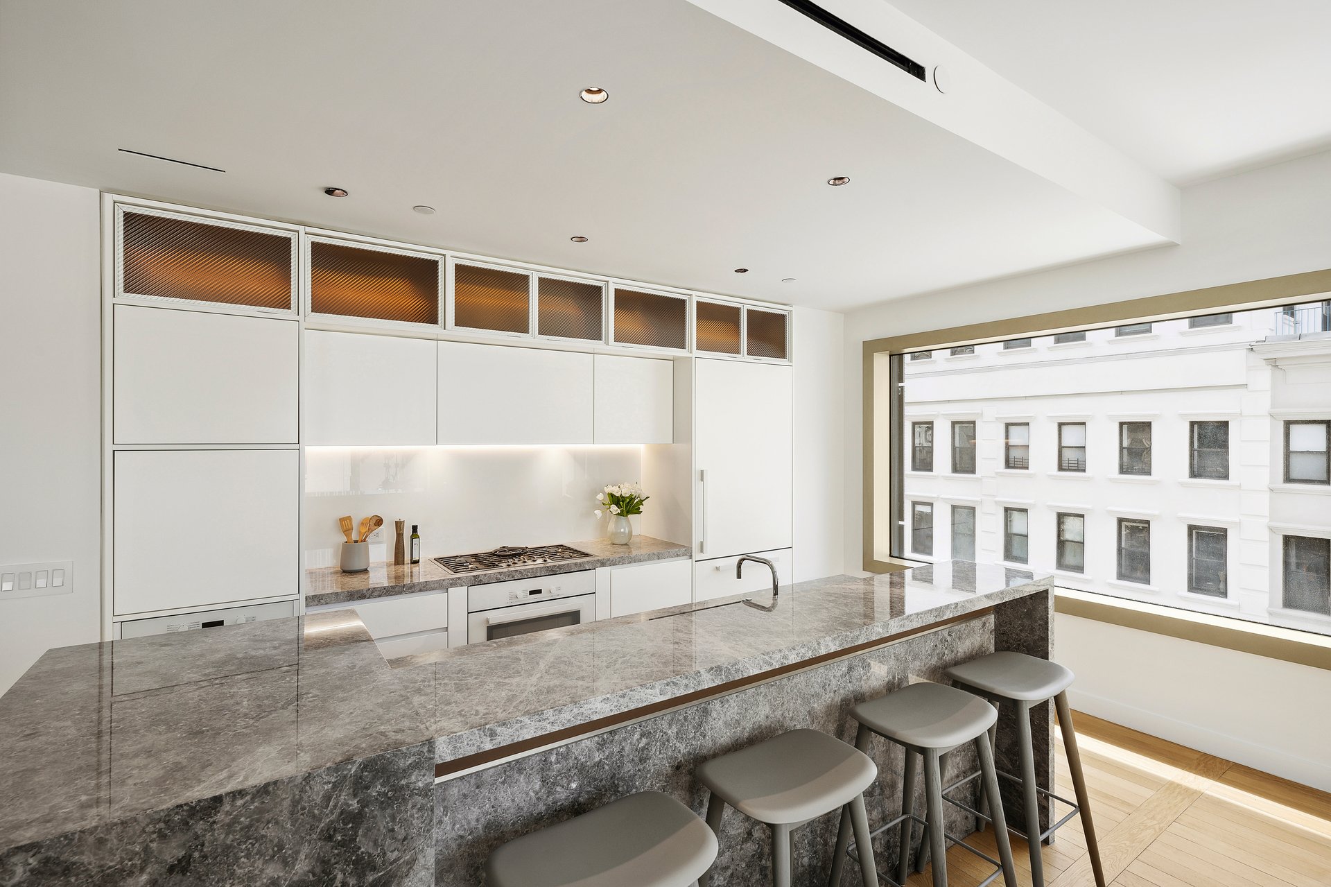 Modern white kitchen with a gray marble island, bar stools, and a large window.