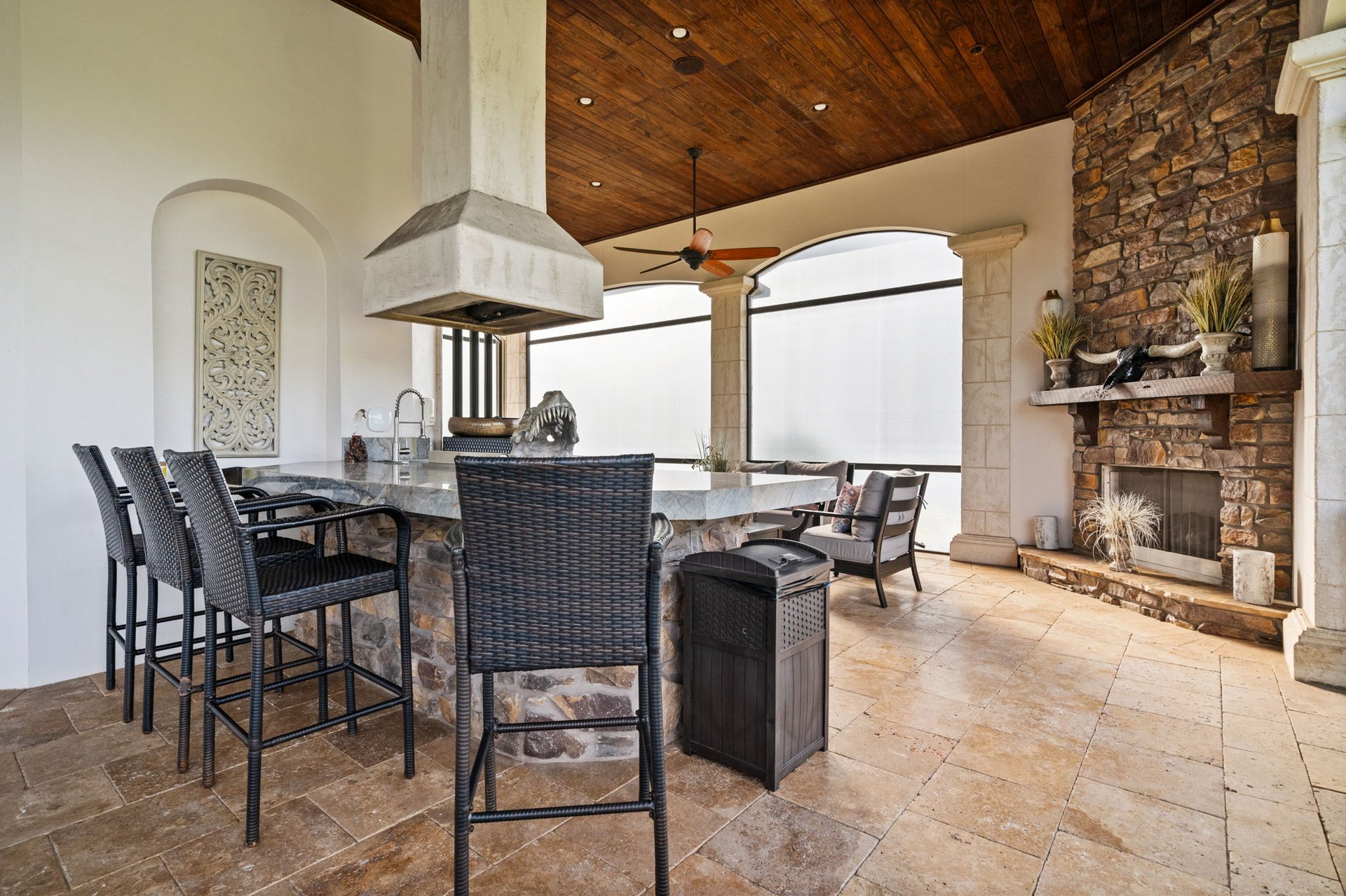 Covered patio with a stone fireplace, built-in bar, woven chairs, and wood-paneled ceiling.
