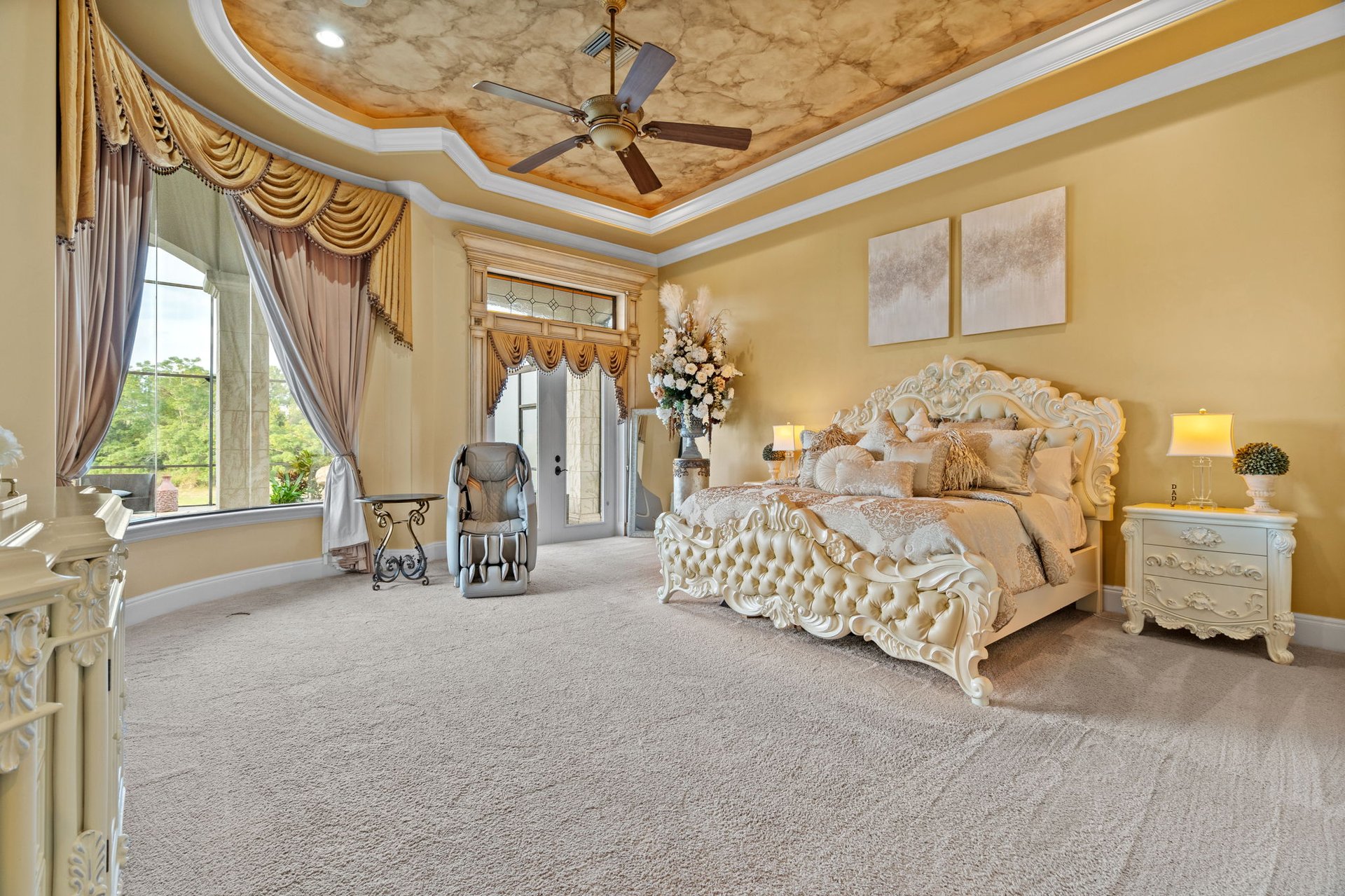 Furnished bedroom with an ornate cream bed, tall draped windows, and a patterned tray ceiling.