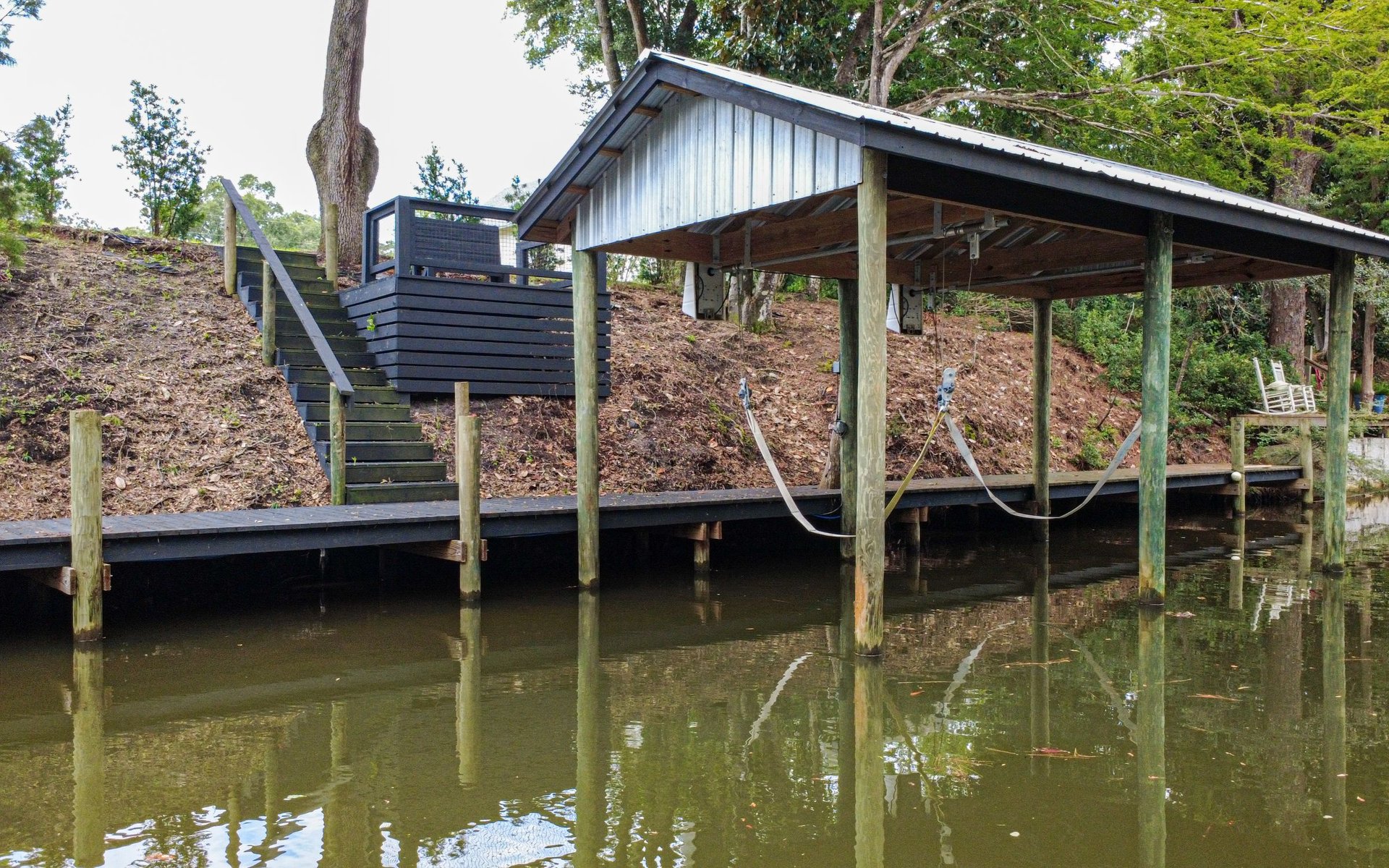 Covered waterfront dock with dark stairs climbing a wooded bank and boat-lift straps over the water.