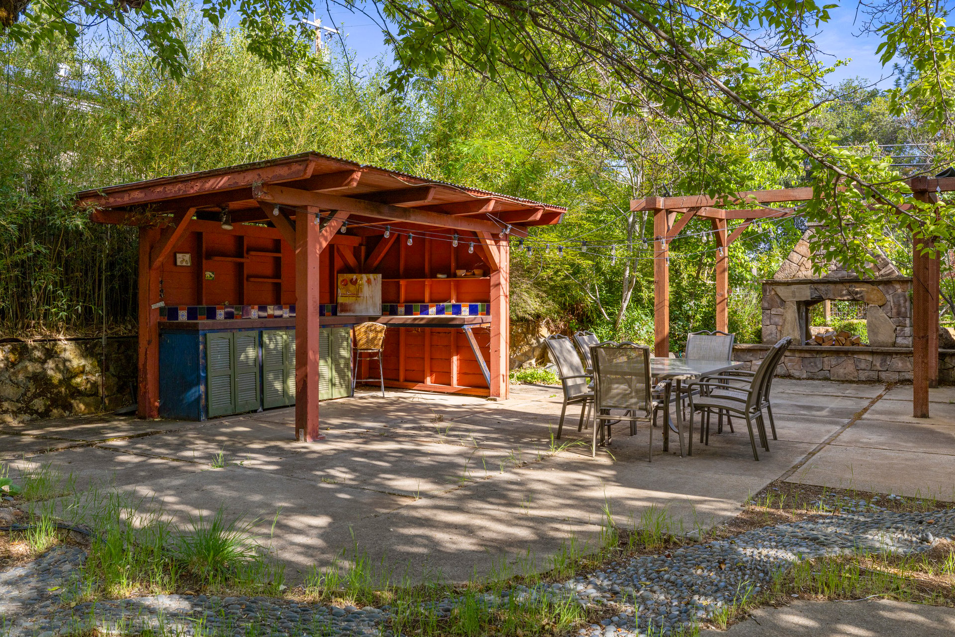 Outdoor patio with a covered kitchen, dining set, pergola, and stone fireplace among leafy trees.