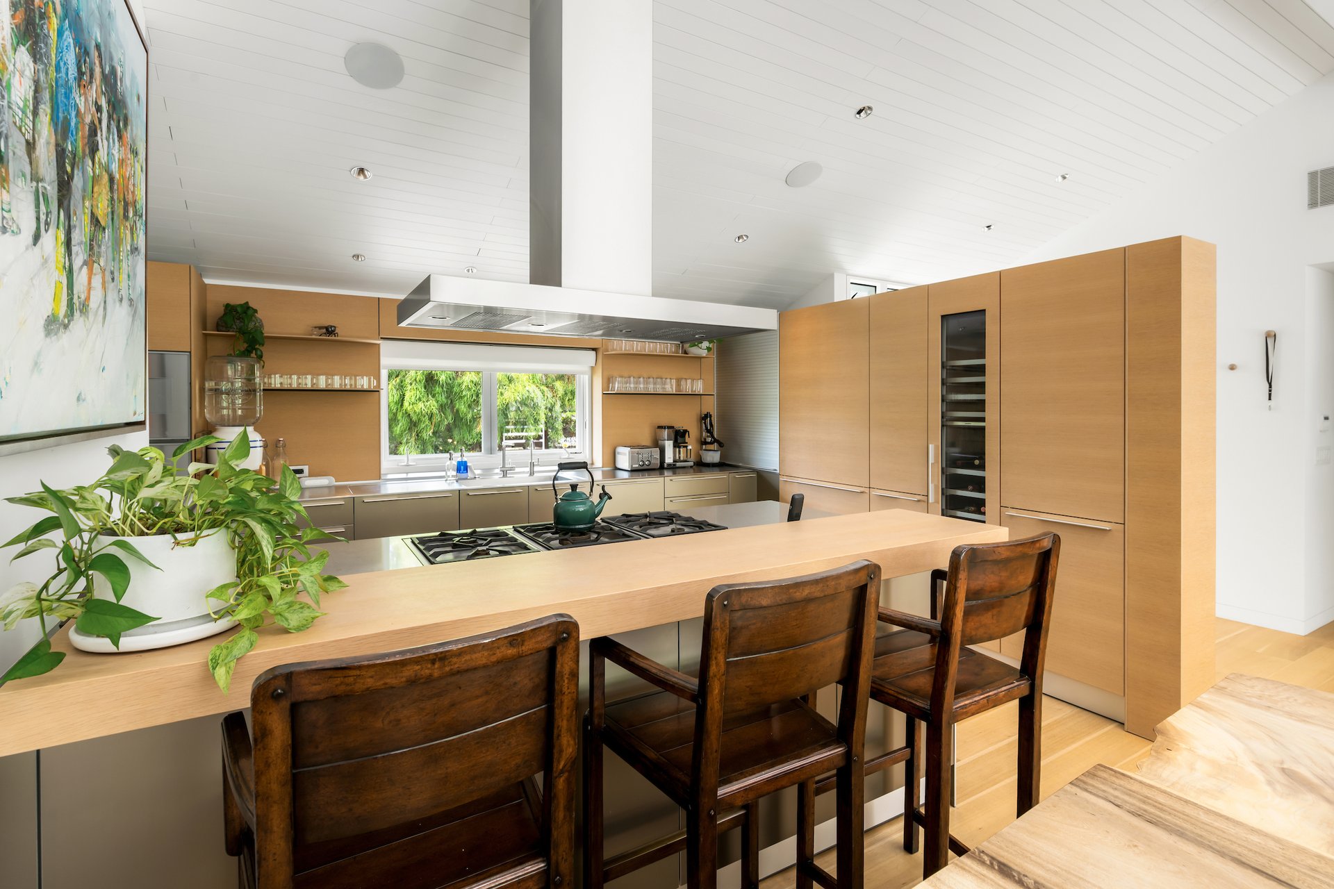 Modern kitchen with light wood cabinetry, a cooktop island, and windows overlooking greenery.