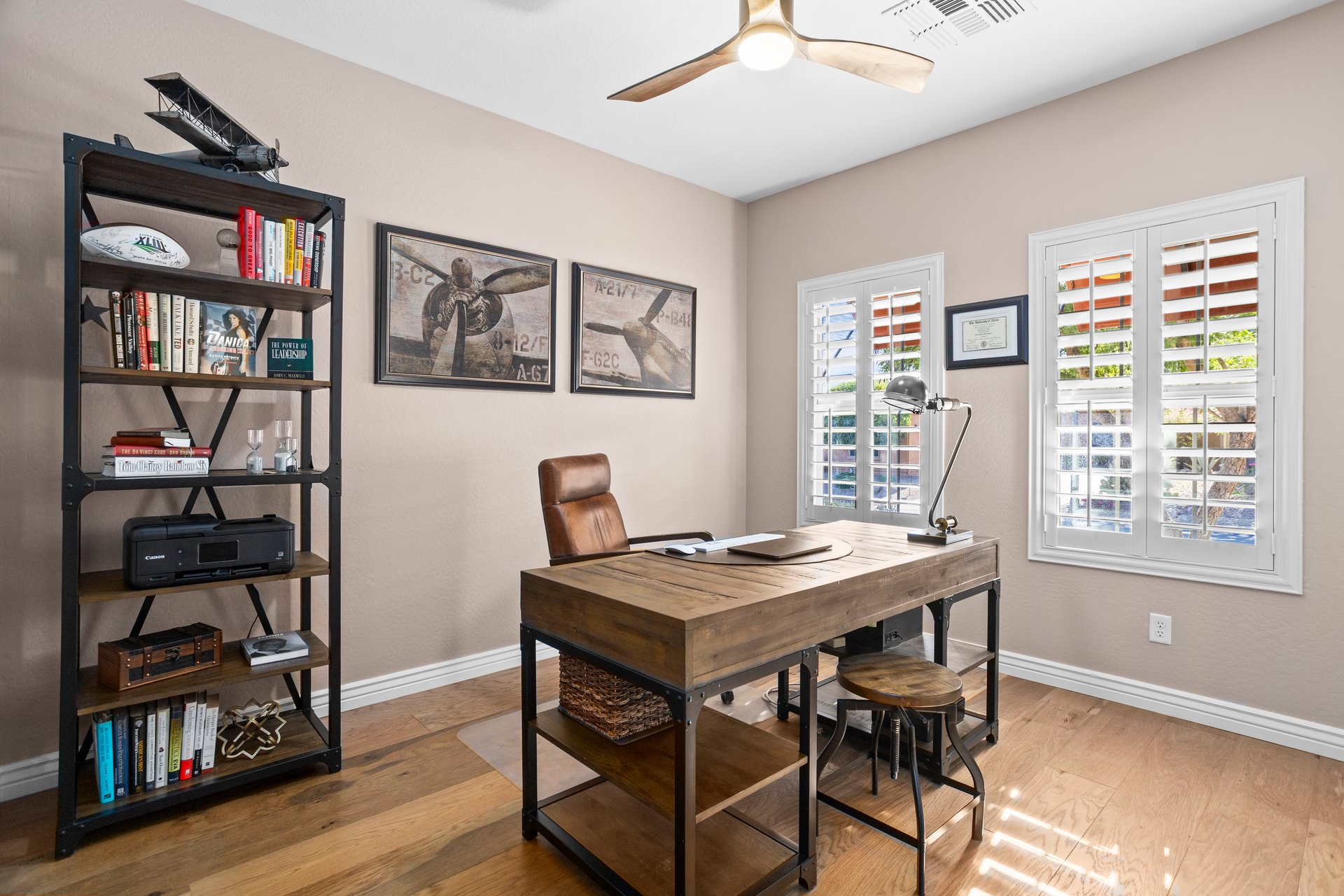 A home office with a wooden desk, bookcase, leather chair, framed airplane art, and shuttered windows.